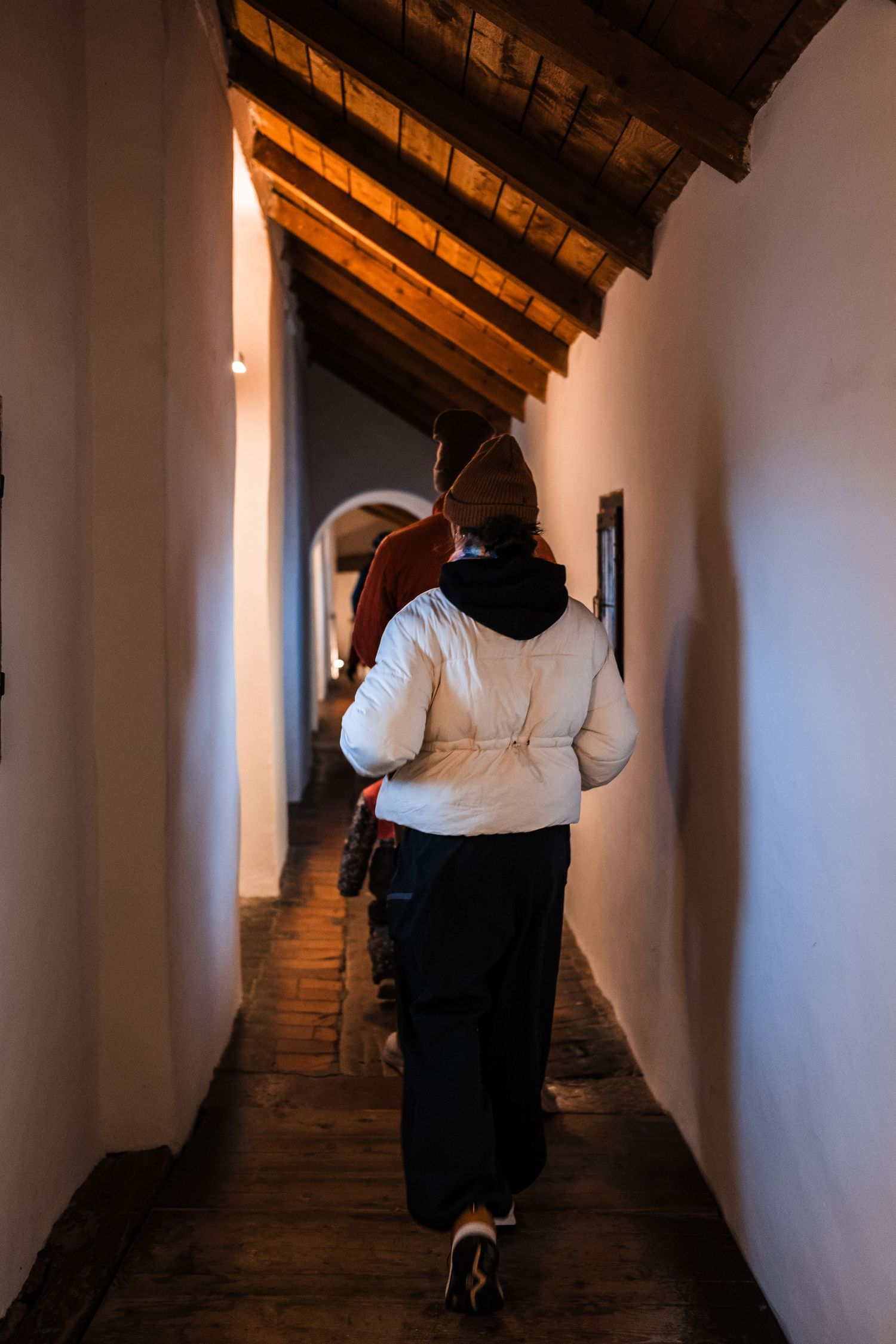 Person walking down dimly lit historic corridor with slanted wooden ceiling and white walls.