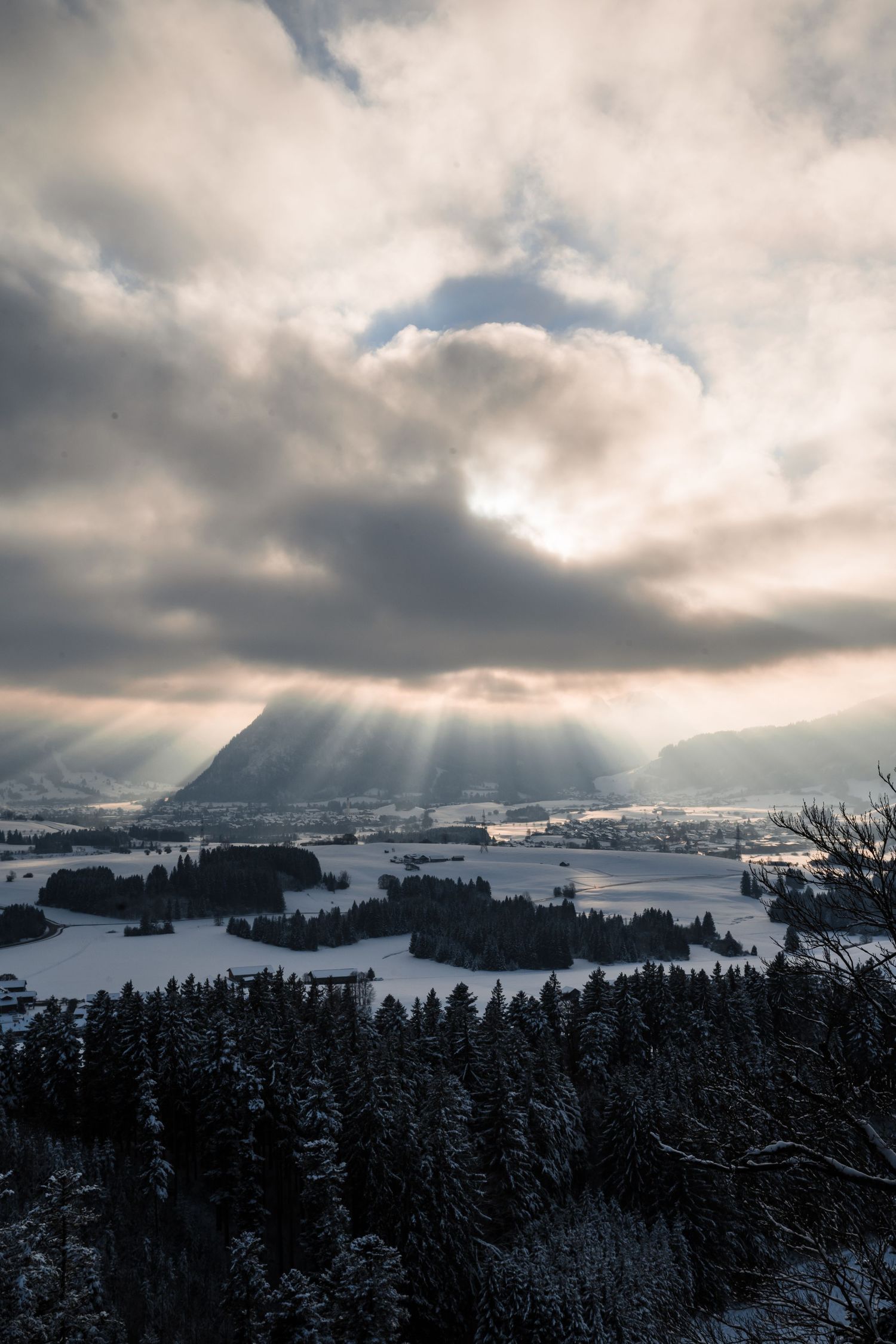 Crepuscular rays shine through clouds onto a snowy mountain valley with evergreen forests.