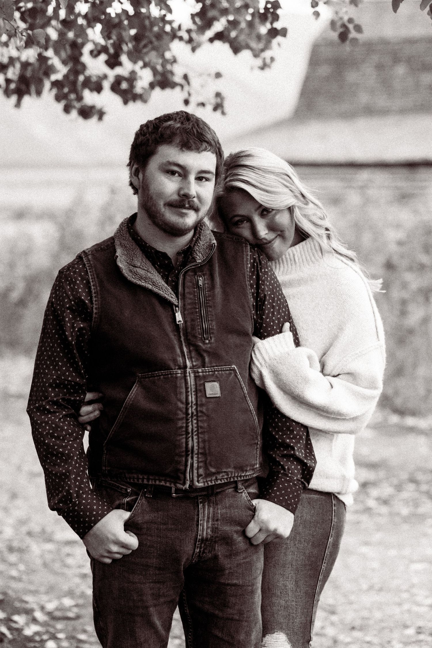 A couple embraces outdoors in autumn, wearing casual clothing including a brown work vest and white sweater.