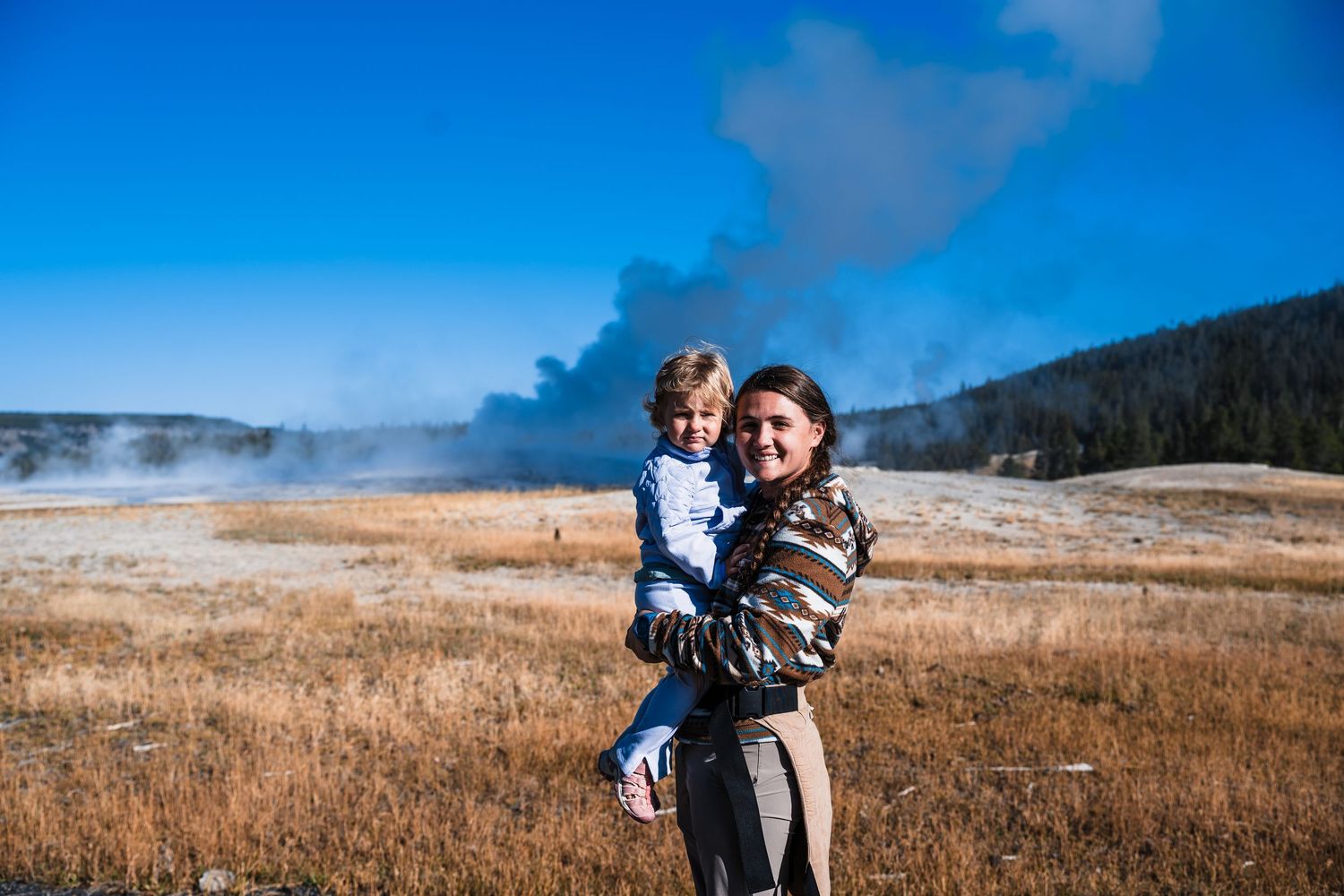 Steam rises from geothermal features in Yellowstone National Park's grassland under bright blue skies.