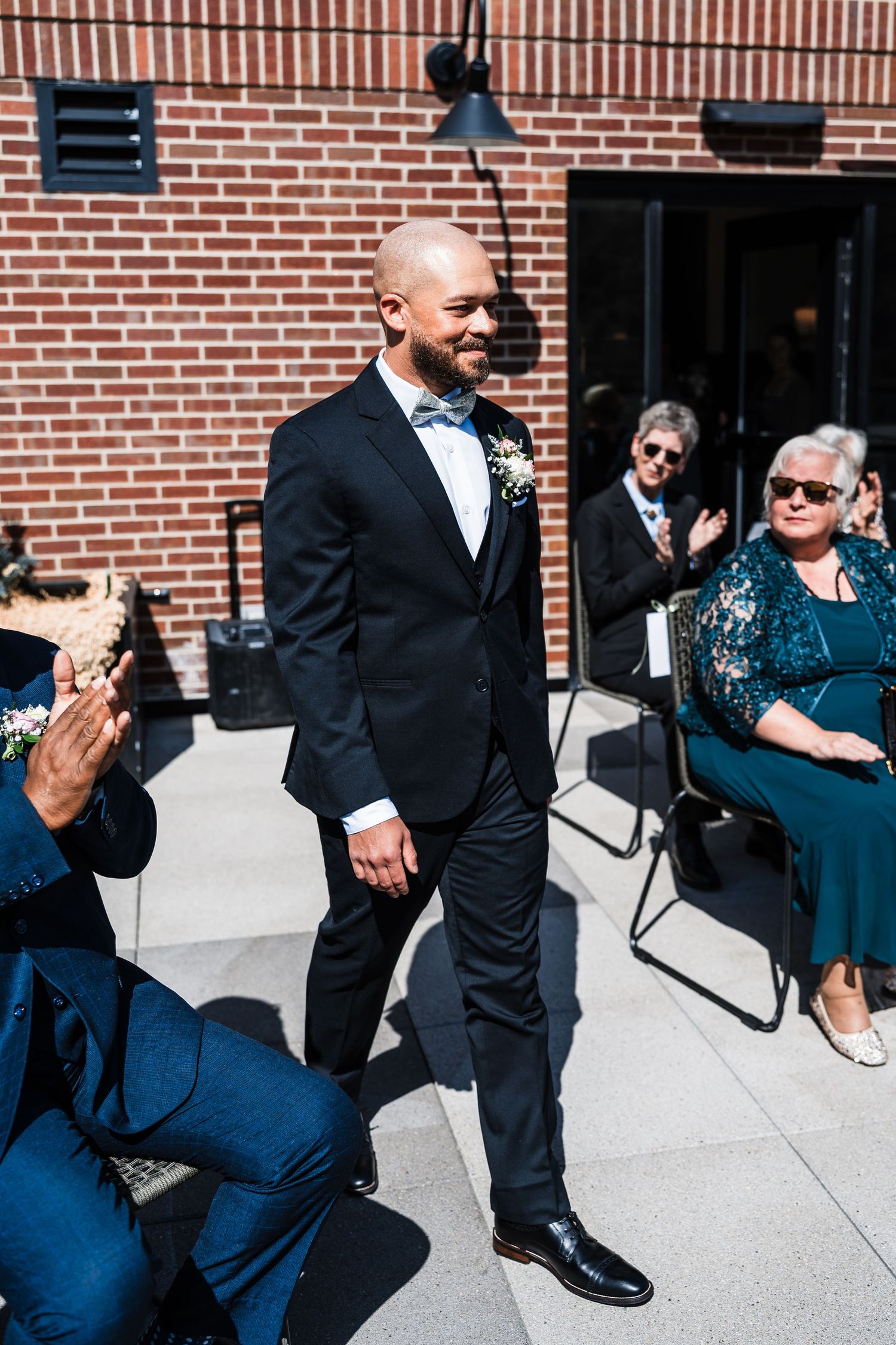 Bald groom in black tuxedo with floral boutonniere walks down aisle as guests applaud.