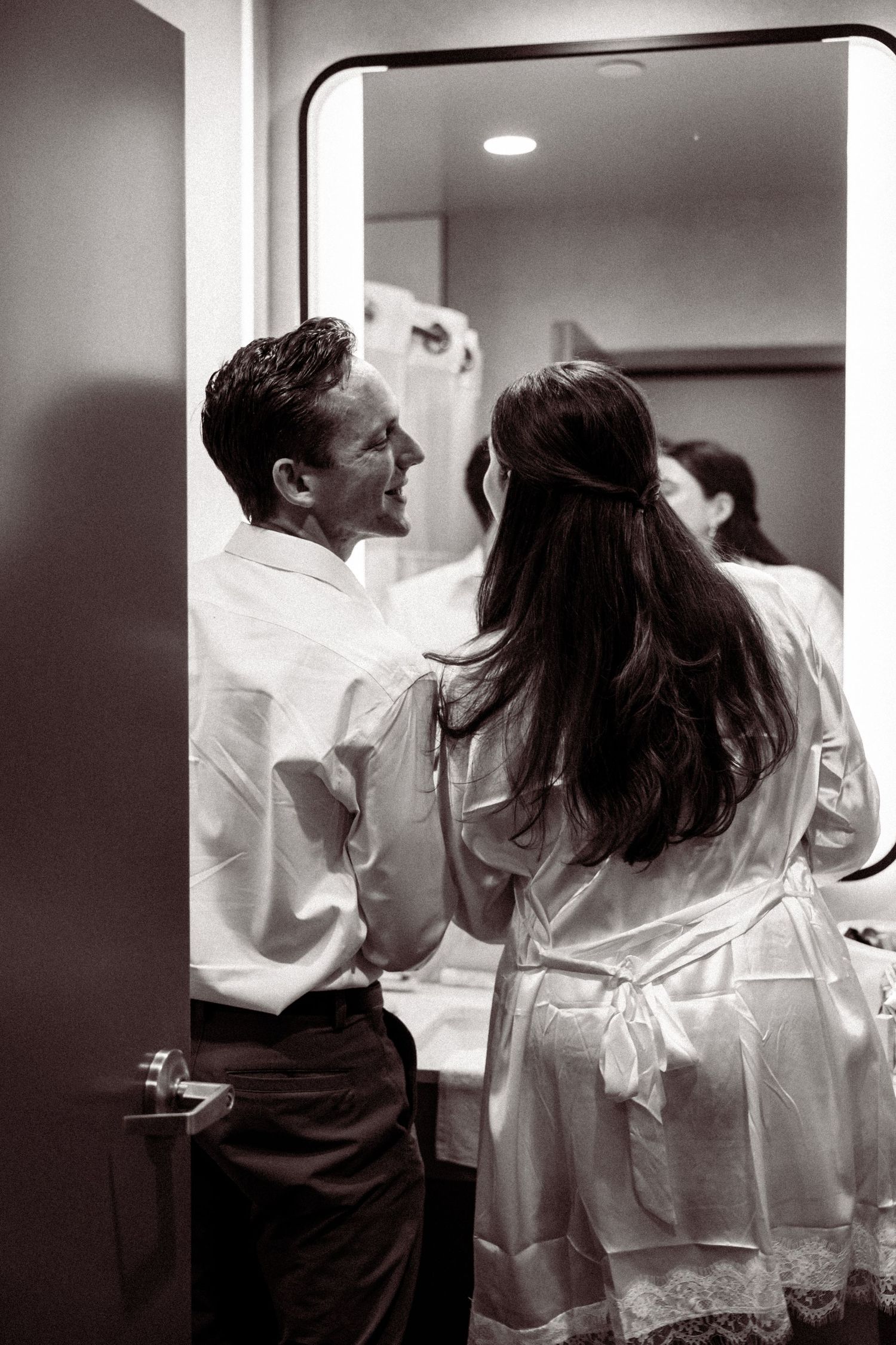 Bride in satin robe helps groom dress before wedding ceremony in black and white photo.