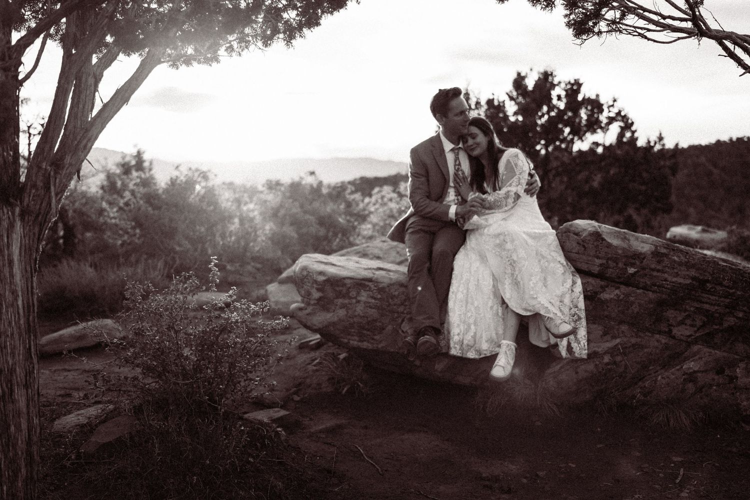 Black and white photo of bride and groom sitting intimately on rocky outcrop surrounded by desert landscape.