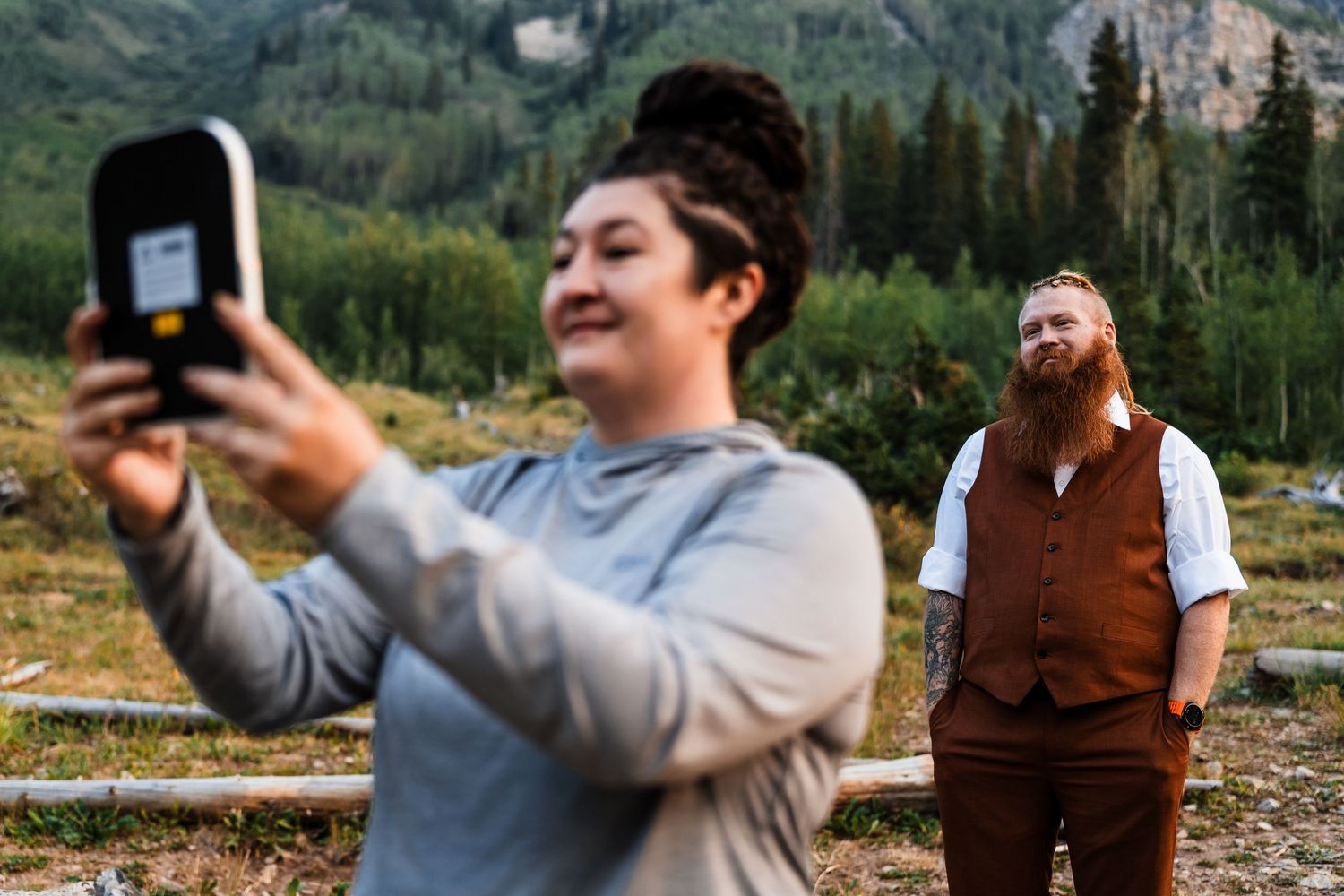 Woman taking selfie outdoors with bearded man in brown vest standing in mountain meadow background.