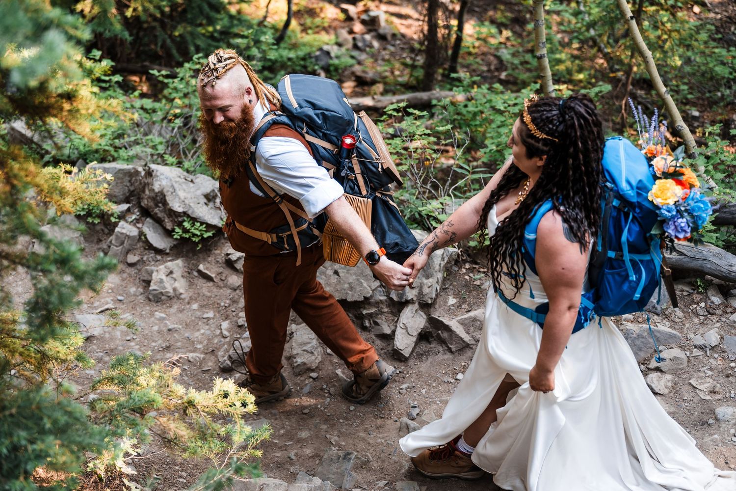 Groom helps bride navigate rocky forest trail while both wear backpacks at adventure elopement.
