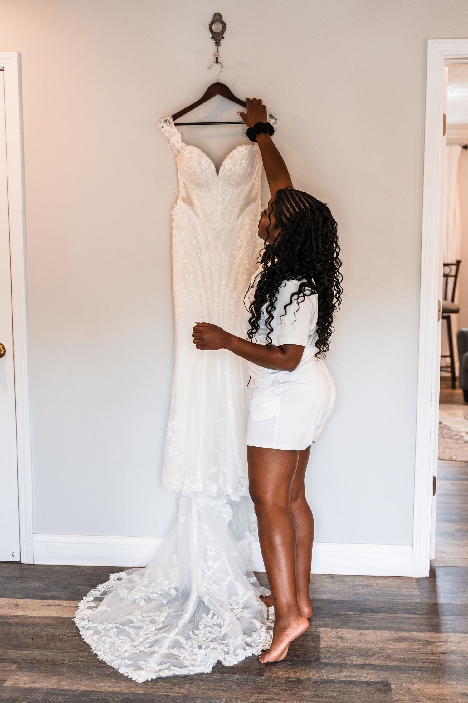 Bride holding her elegant beaded white wedding gown with lace train on a hanger against a neutral wall.