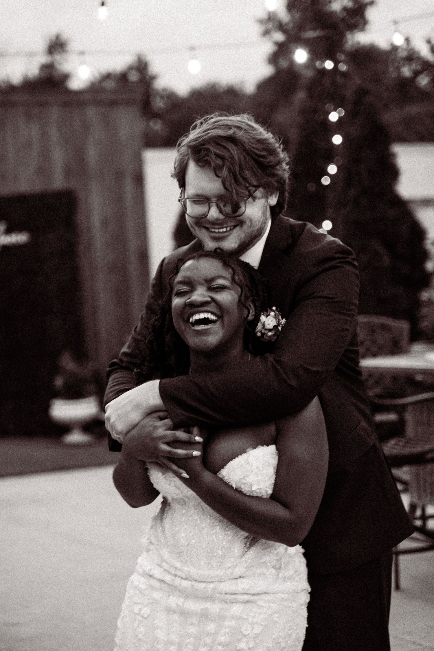 Groom hugs laughing bride from behind in joyful black and white outdoor reception photo.