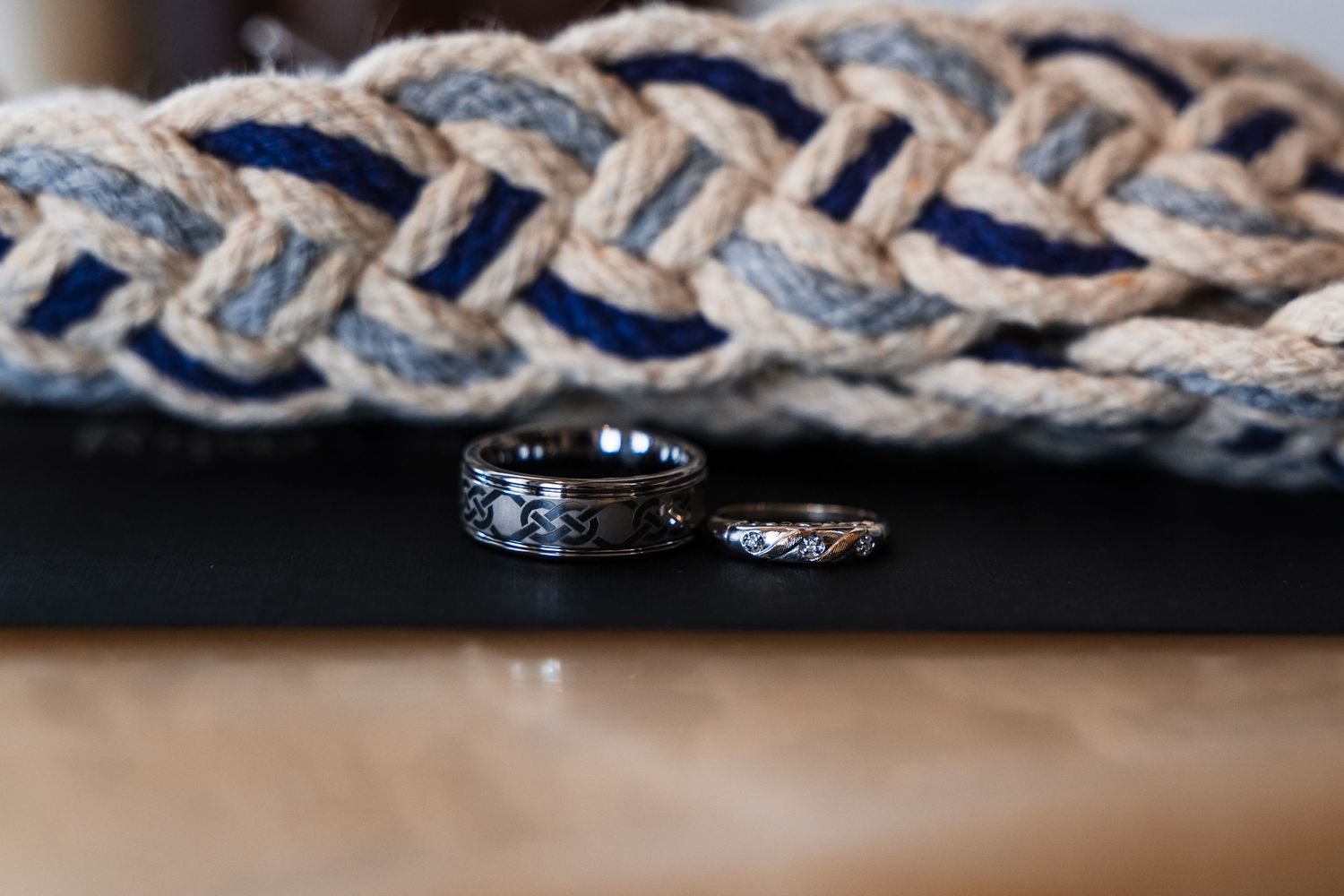 Two black wedding bands with intricate designs displayed on a dark surface beside a braided rope backdrop.