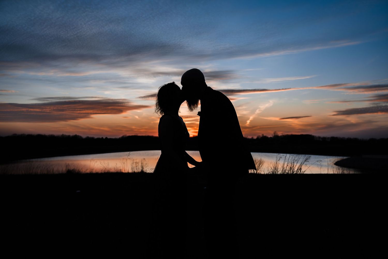 Romantic silhouette of couple sharing kiss by lake at stunning colorful sunset during wedding.