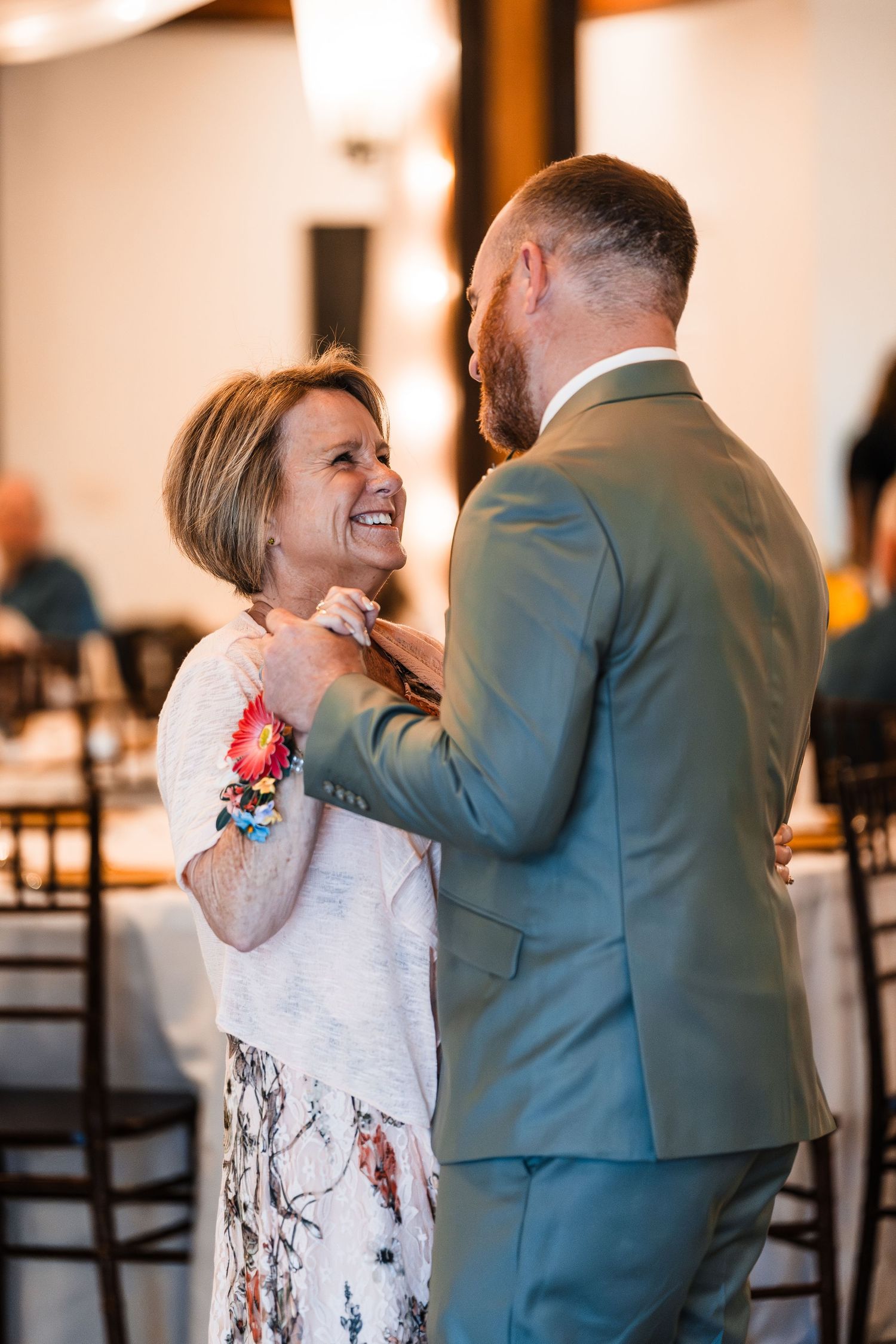 Groom in sage suit shares a joyful mother-son dance at warmly lit wedding reception.