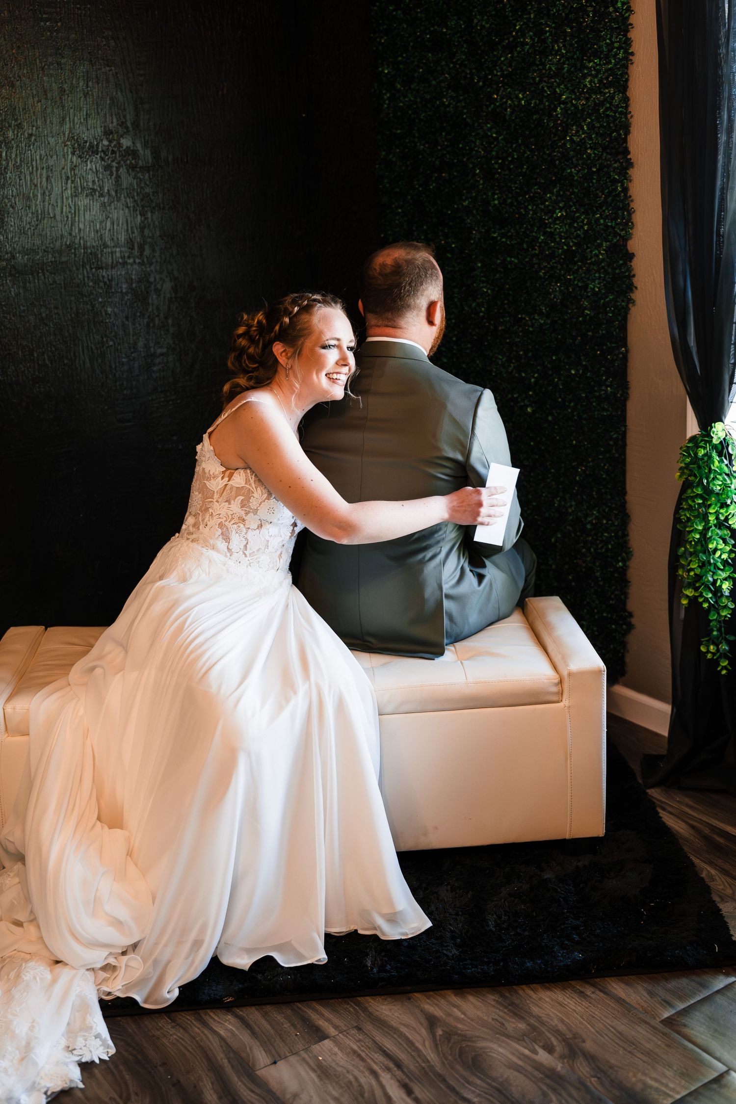 Bride in white lace gown laughing while sitting back-to-back with groom on white bench before wedding ceremony.