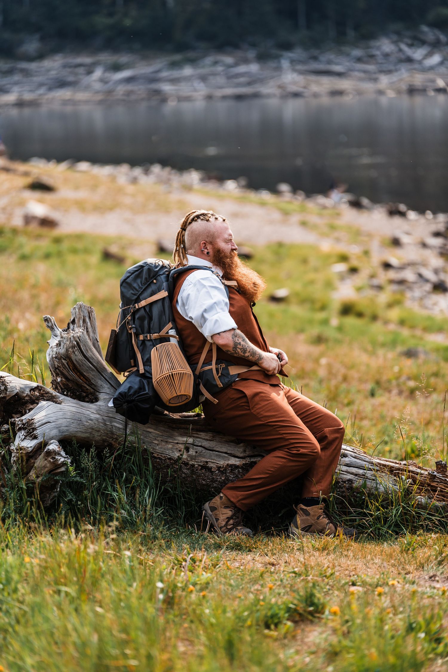 A bearded red-haired man with dreadlocks and a backpack rests on a log by a scenic mountain river.