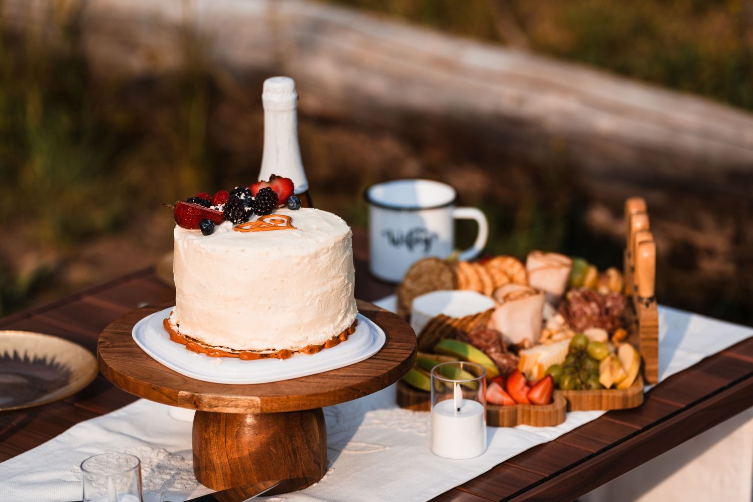 White wedding cake with berries on wooden stand beside charcuterie board at outdoor elopement picnic.