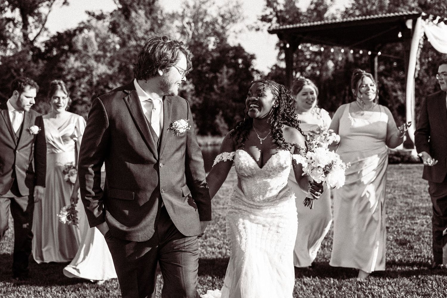 Black and white photo of newlyweds walking with wedding party after outdoor ceremony.