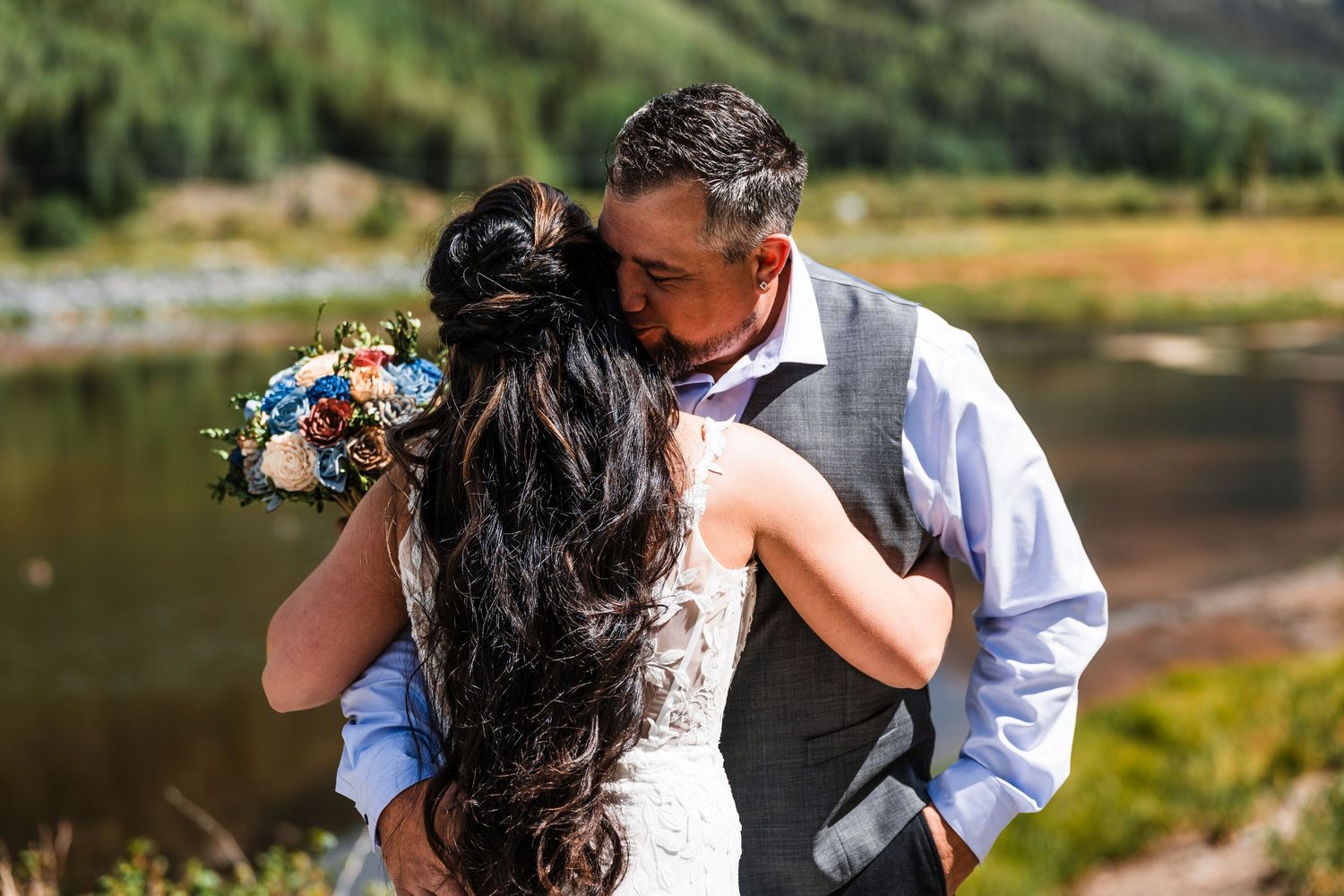 Bride and groom share a tender kiss outdoors near a mountain lake, bride holding a colorful bouquet.