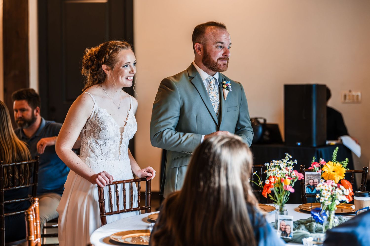 Smiling bride in lace gown and groom in gray suit greet guests at wedding reception.