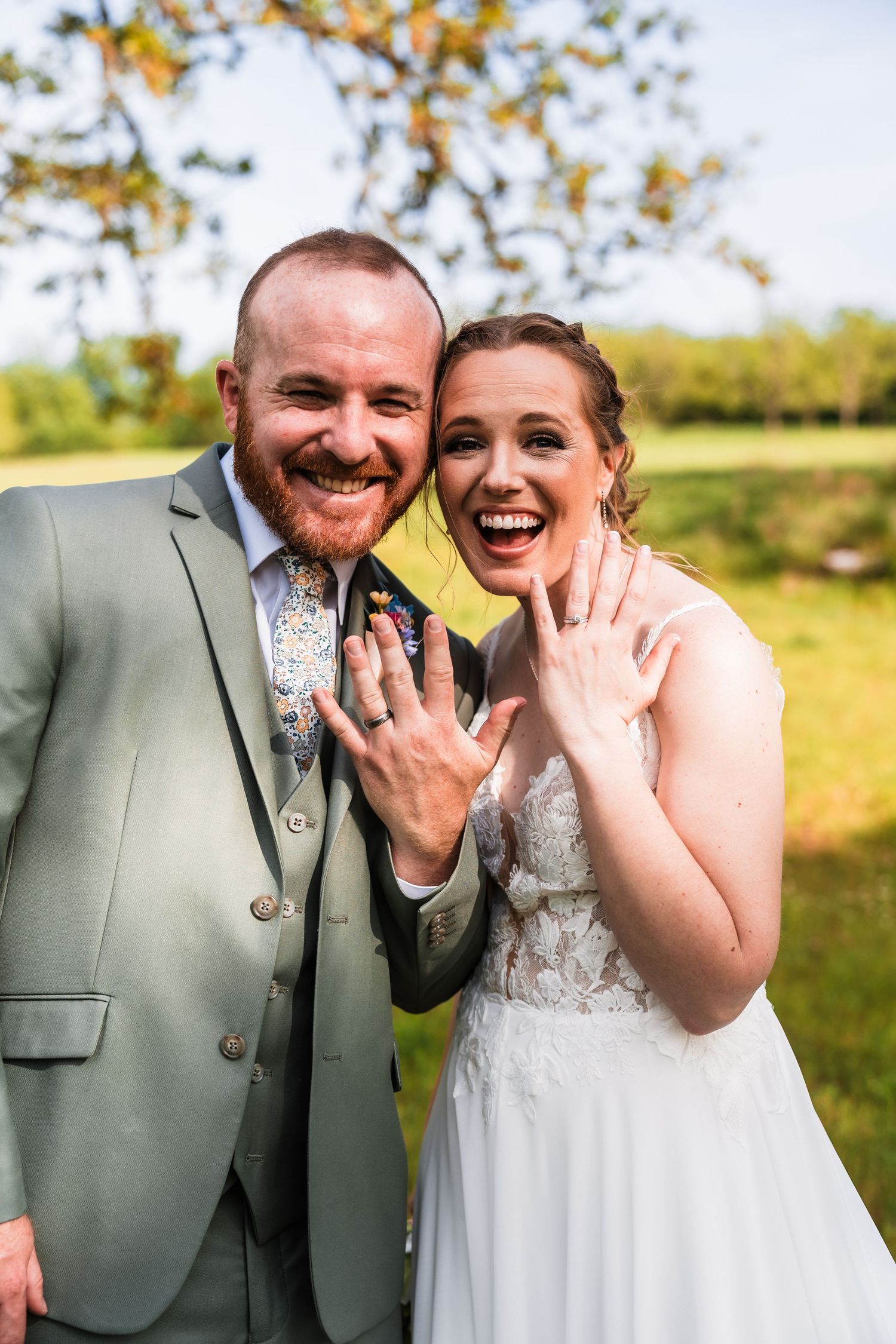 Joyful newlywed couple smiling and showing off wedding rings outdoors on sunny wedding day.