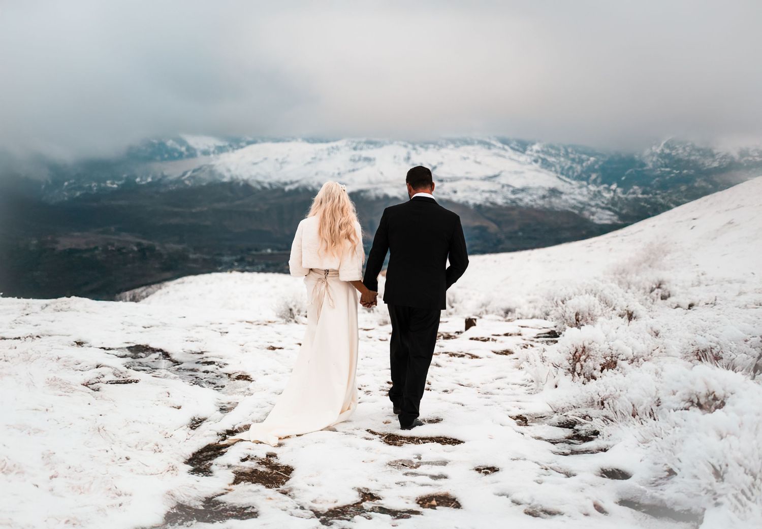 Bride and groom walking hand in hand on snowy mountain summit.