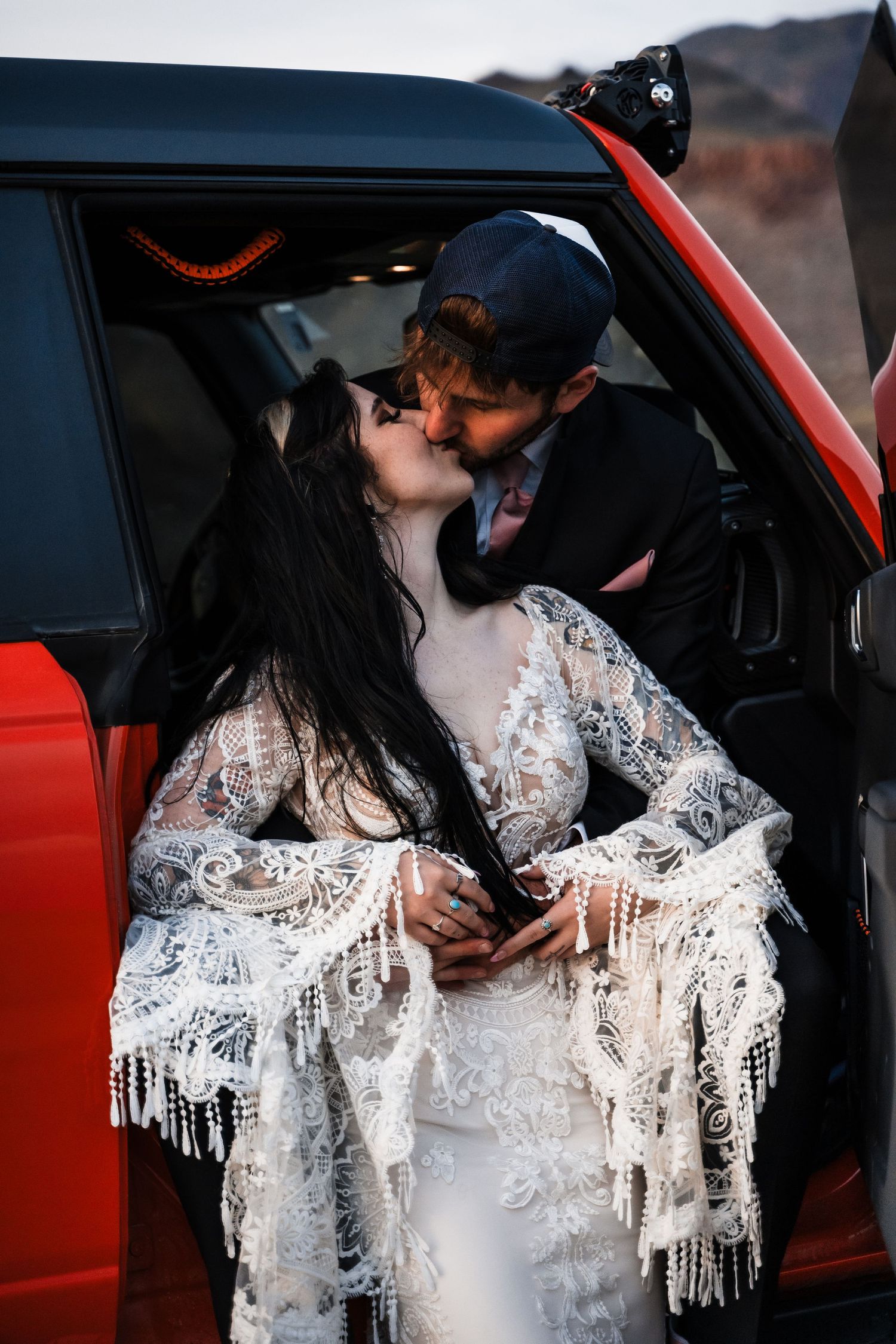 A romantic moment captured inside a red vehicle with the bride in a detailed lace gown.