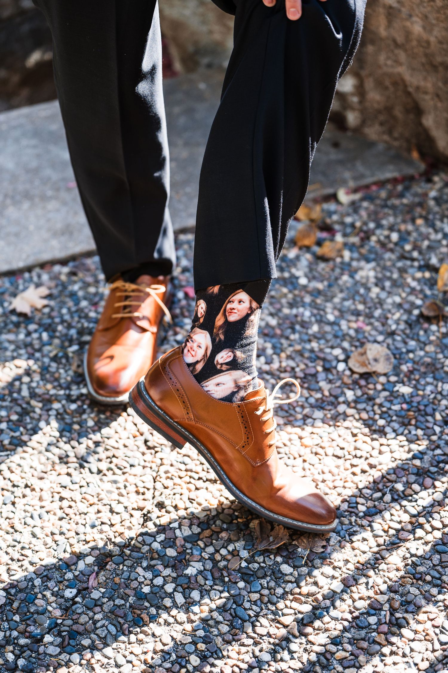 Close-up of groom's brown leather dress shoes and unique patterned socks on gravel.