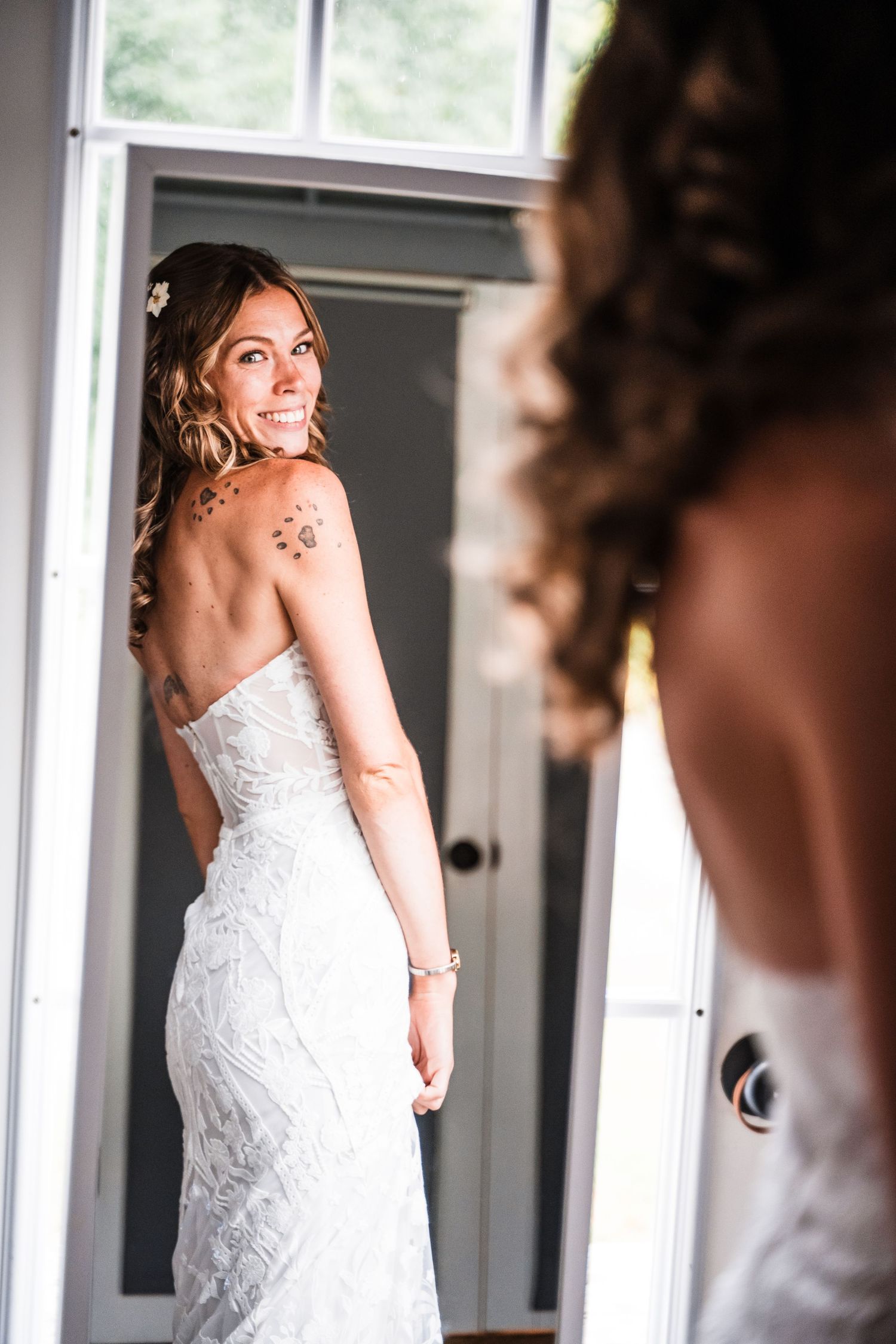 Bride shows off detailed lace wedding dress while looking over shoulder in mirror.