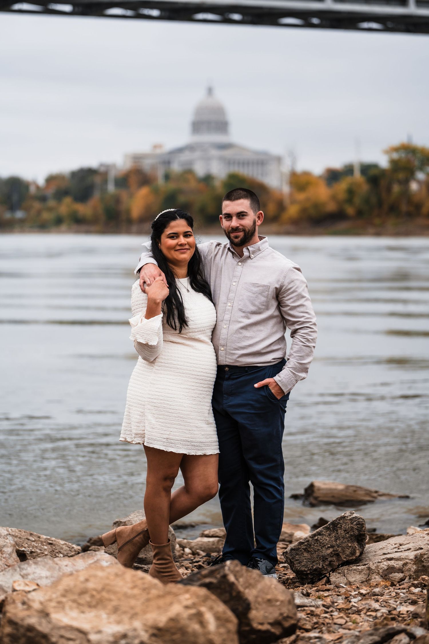 Couple posing by rocky shoreline with state capitol dome visible across water.