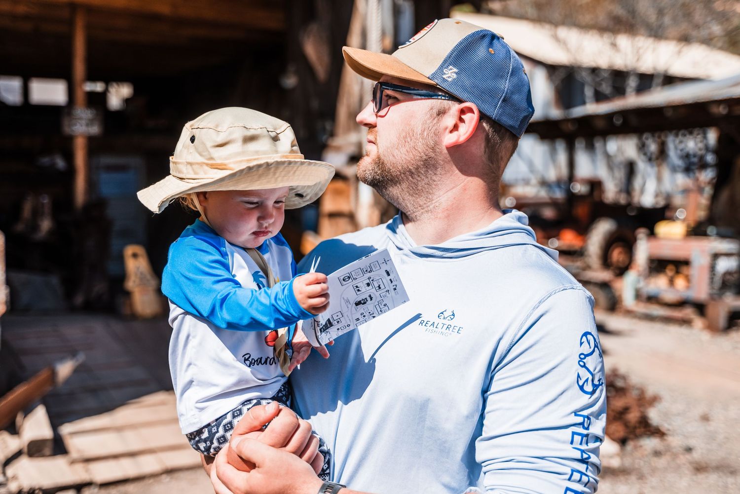 Outdoor scene showing someone holding a small child wearing a sun hat against a rustic backdrop.