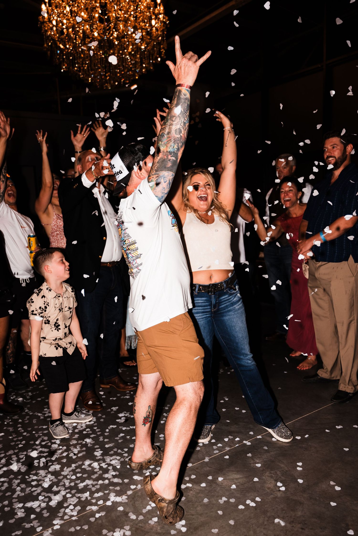 Happy party crowd dancing under a chandelier as confetti falls during a celebratory event.