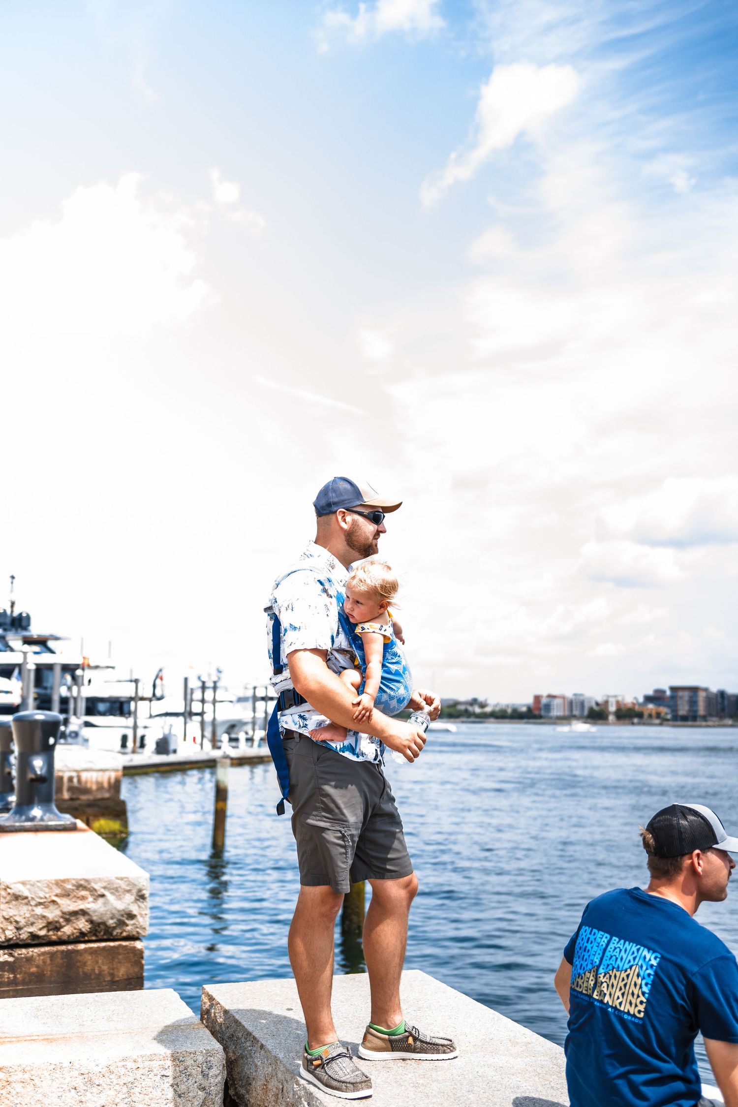 People standing on a pier overlooking a marina and waterfront on a sunny day.