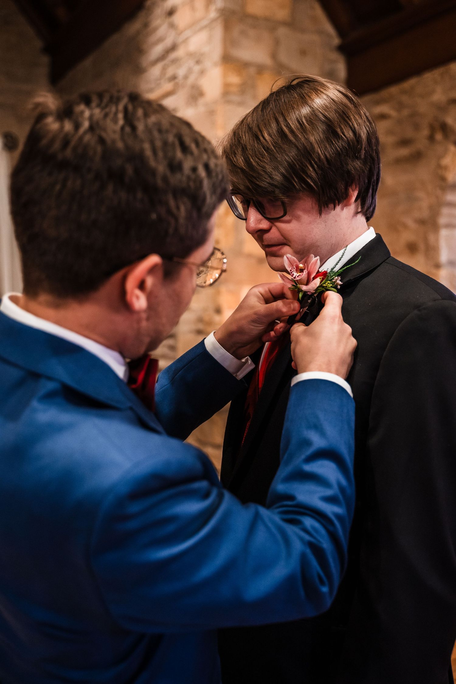 A groomsman in a blue suit helps adjust another's red tie and boutonniere before the wedding ceremony.