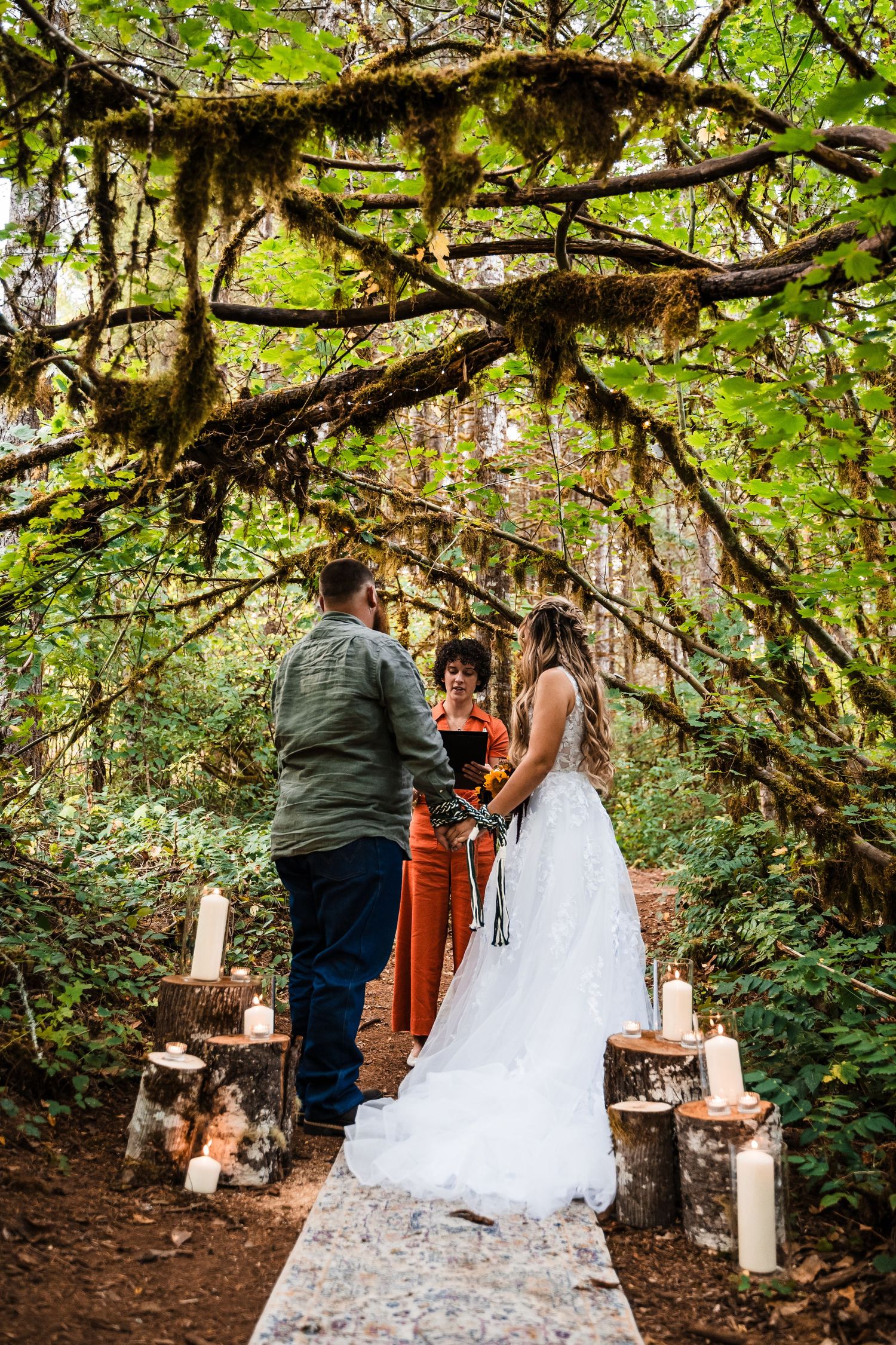 Intimate woodland wedding ceremony with white runner and lit candles on natural log pedestals.
