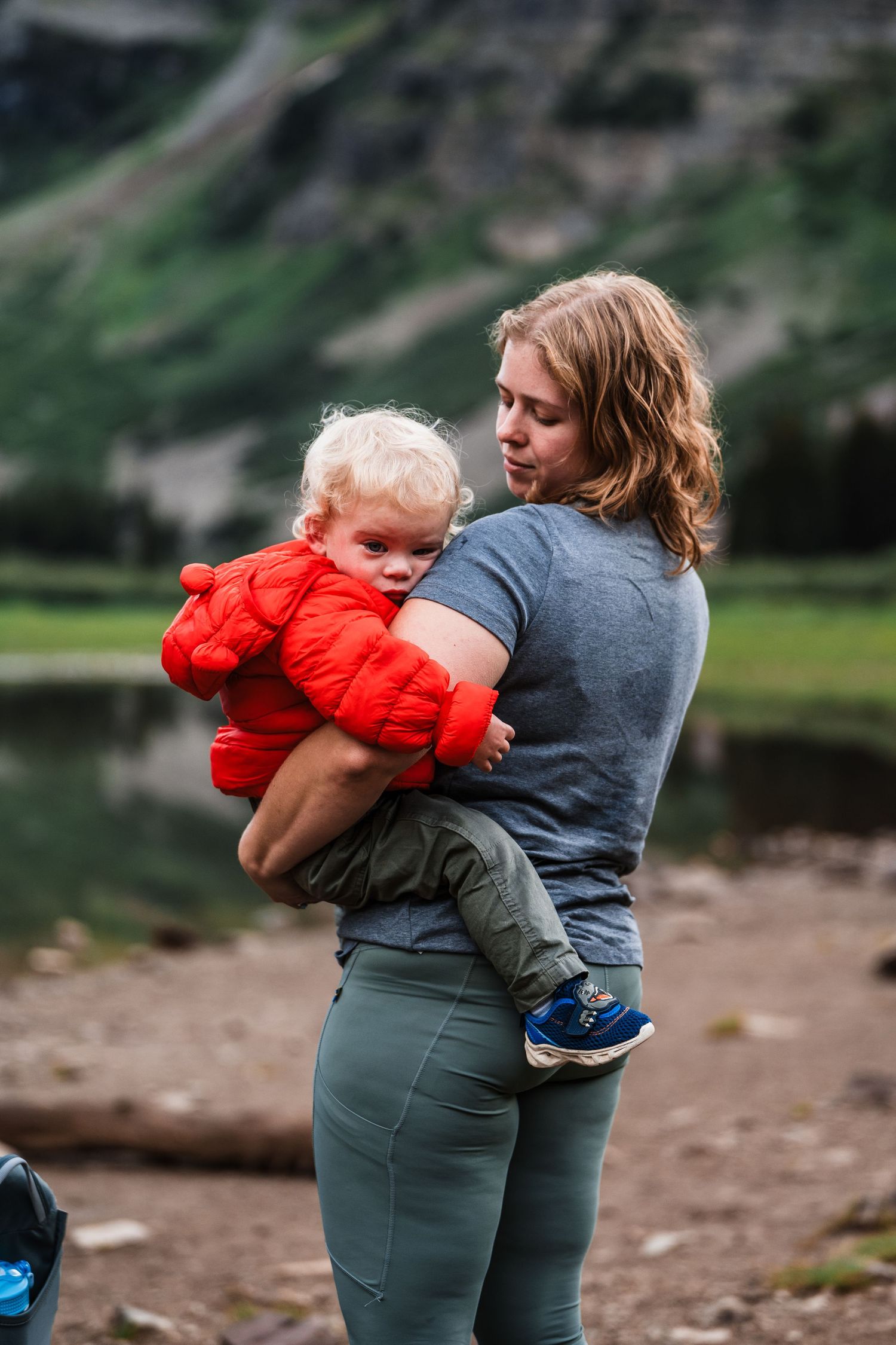 Adult holding child in bright red jacket by peaceful mountain lake.