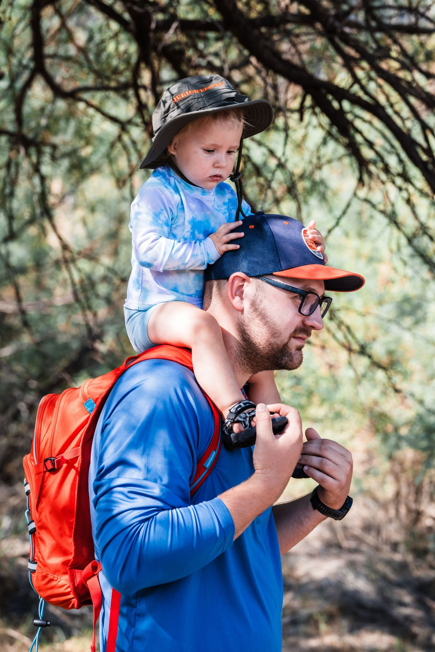 Hiker in blue jacket with orange backpack carries a small child in a hat on their shoulders during a nature walk.