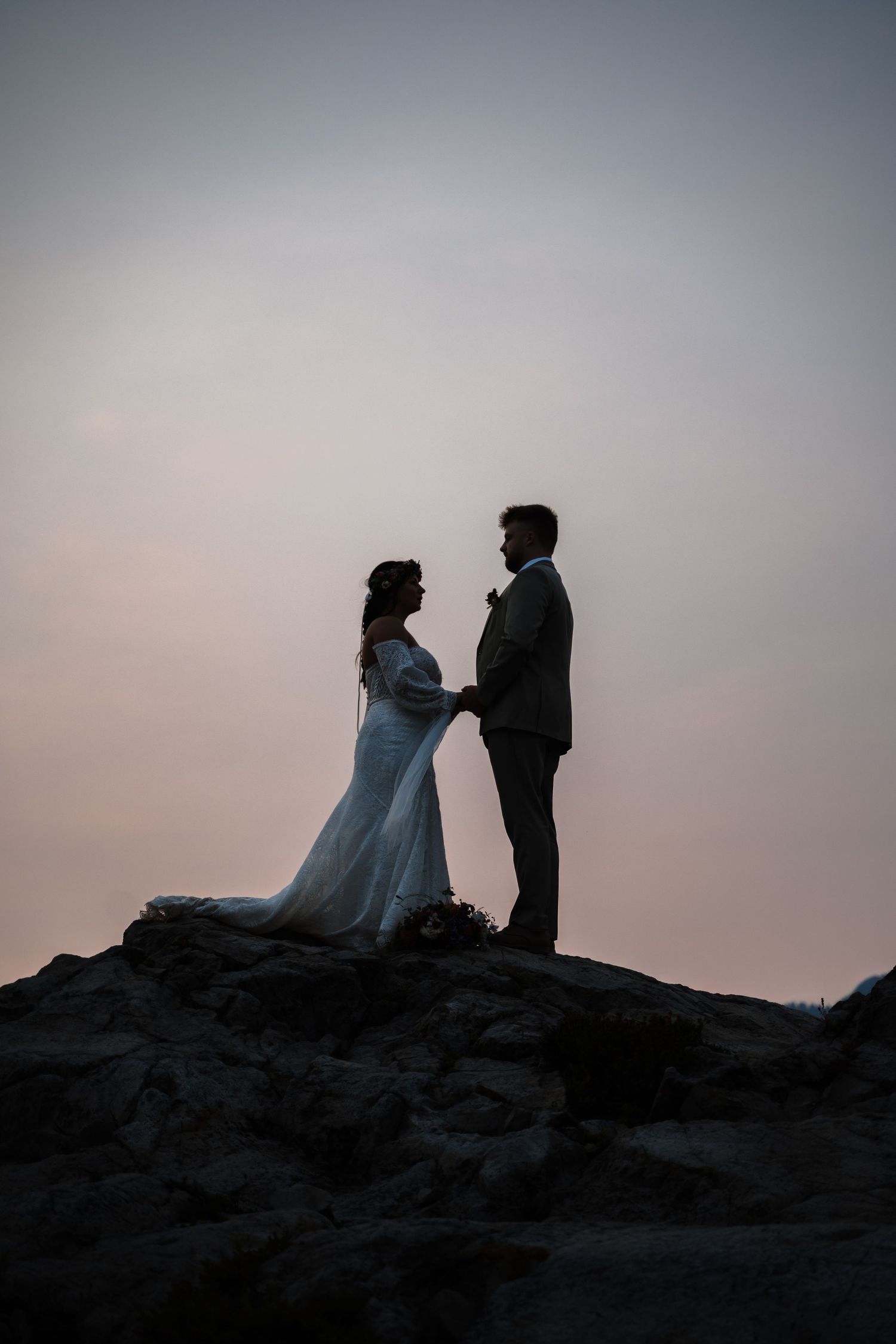 Silhouettes of a couple in wedding attire stand dramatically against a dusky sky on rocky terrain.