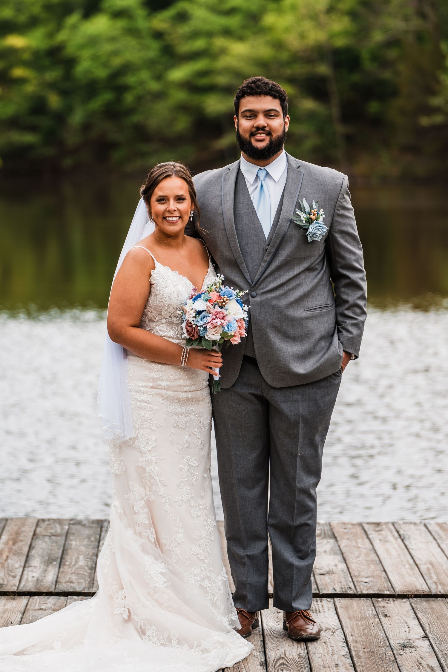 Wedding couple poses on wooden dock overlooking lake with floral bouquet.