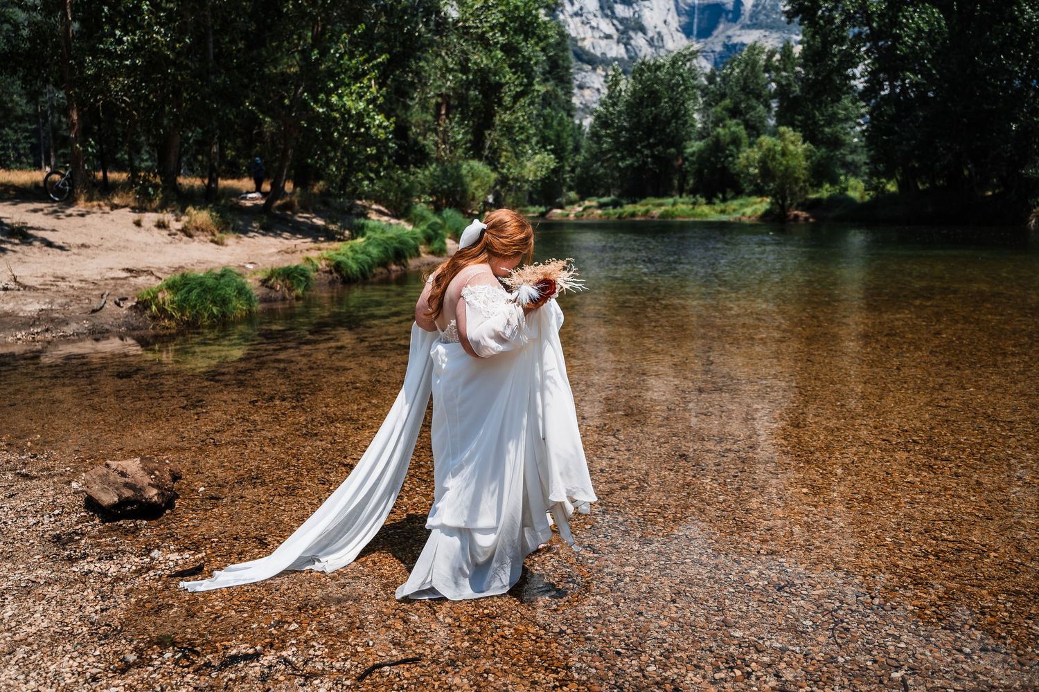 A bride in a flowing white dress and cape stands on the shore of a serene river surrounded by green trees and forest.