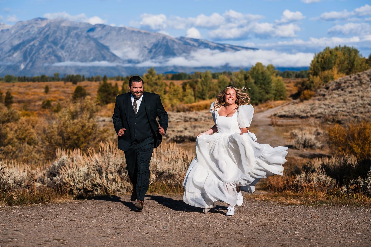 A couple in wedding attire sits together against a backdrop of rugged mountains and autumn foliage.