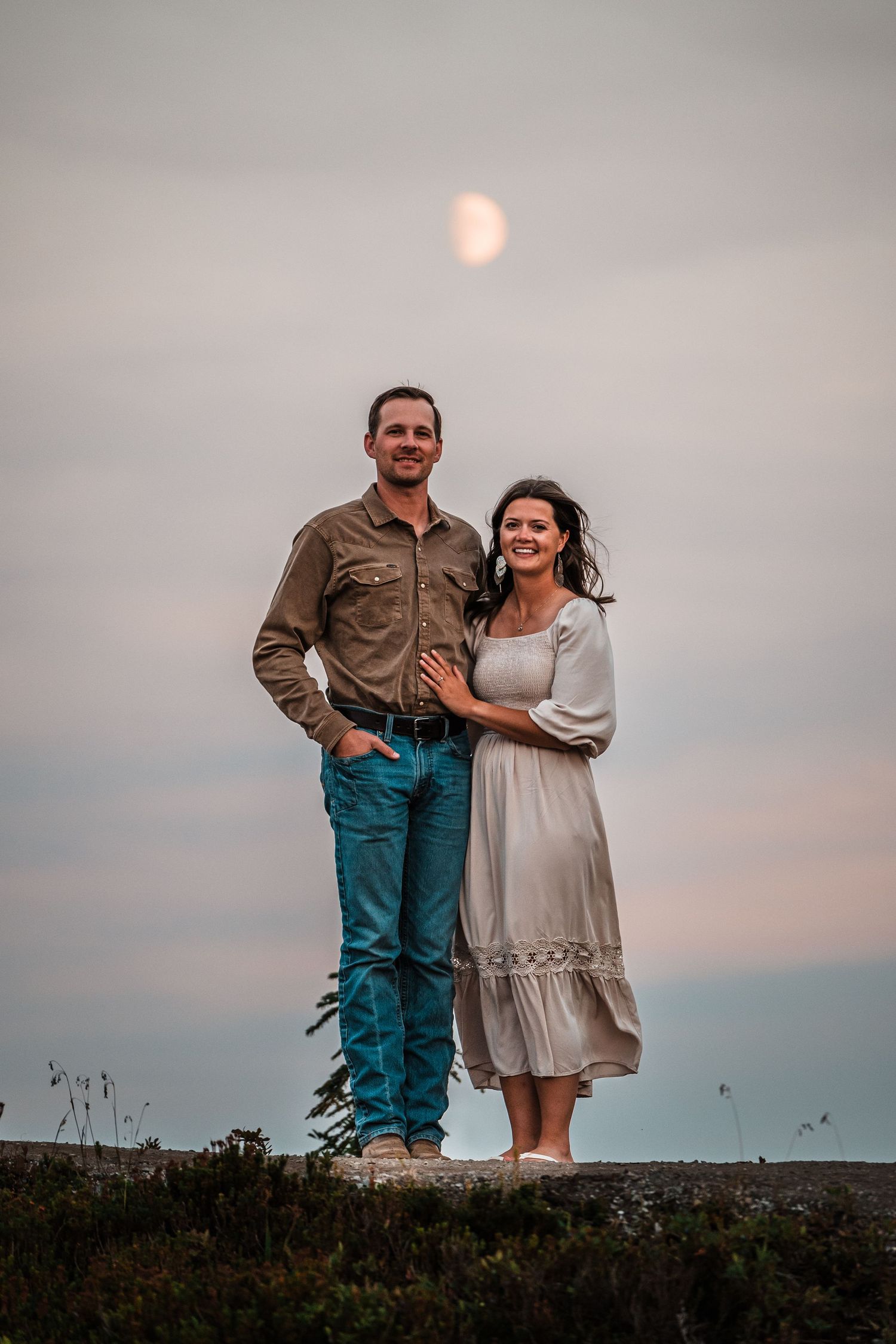 A couple stands together on a hilltop with a half moon visible in the dusky sky behind them.