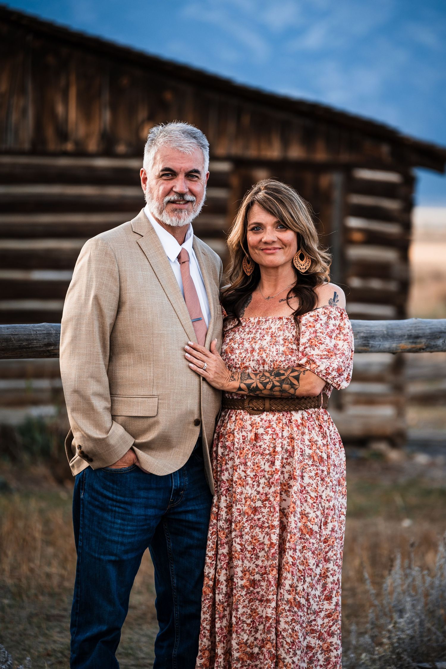 A couple poses together outdoors near a rustic wooden barn in a scenic country setting at sunset.