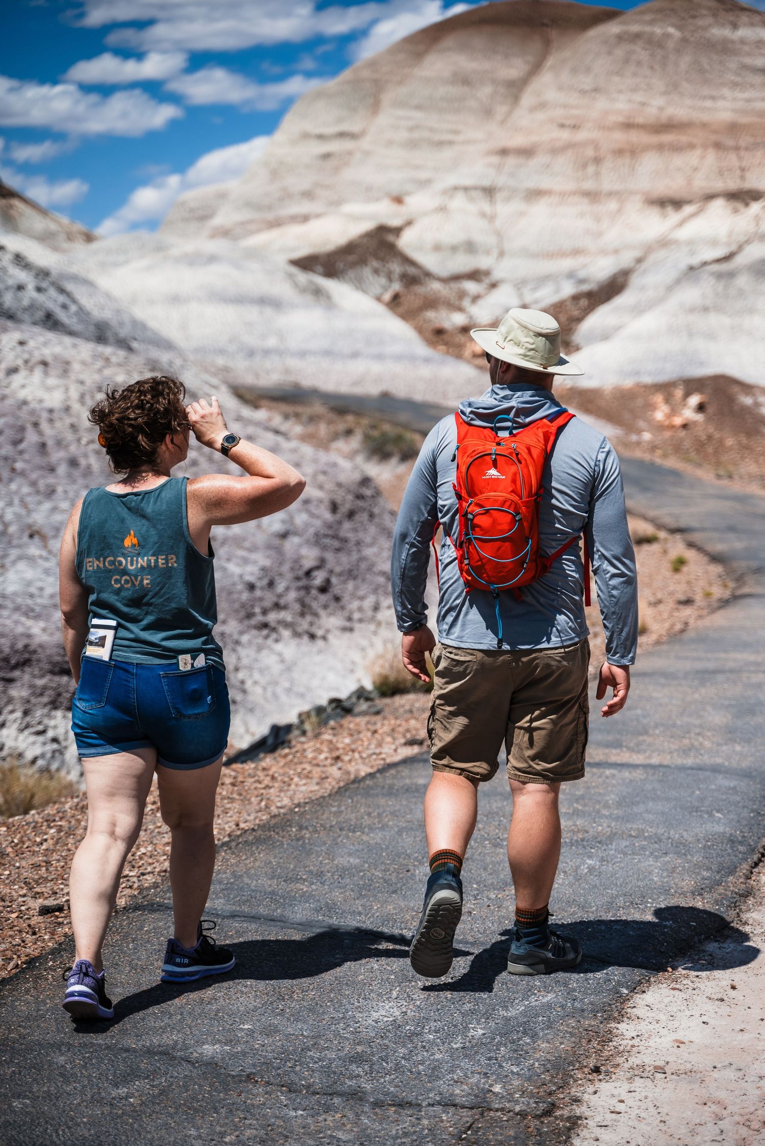 Two hikers walking on a paved path through dramatic striped desert rock formations.