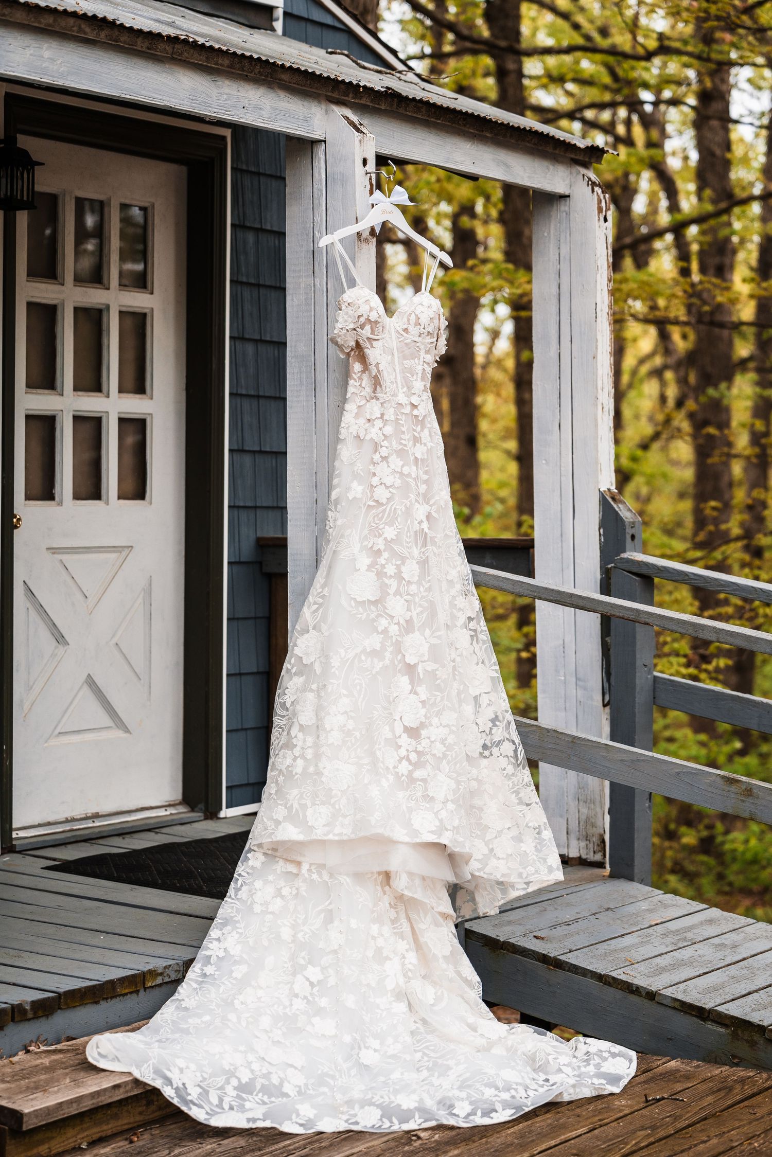 A delicate white lace wedding dress with a long train hangs on display on a rustic cabin porch surrounded by autumn trees.