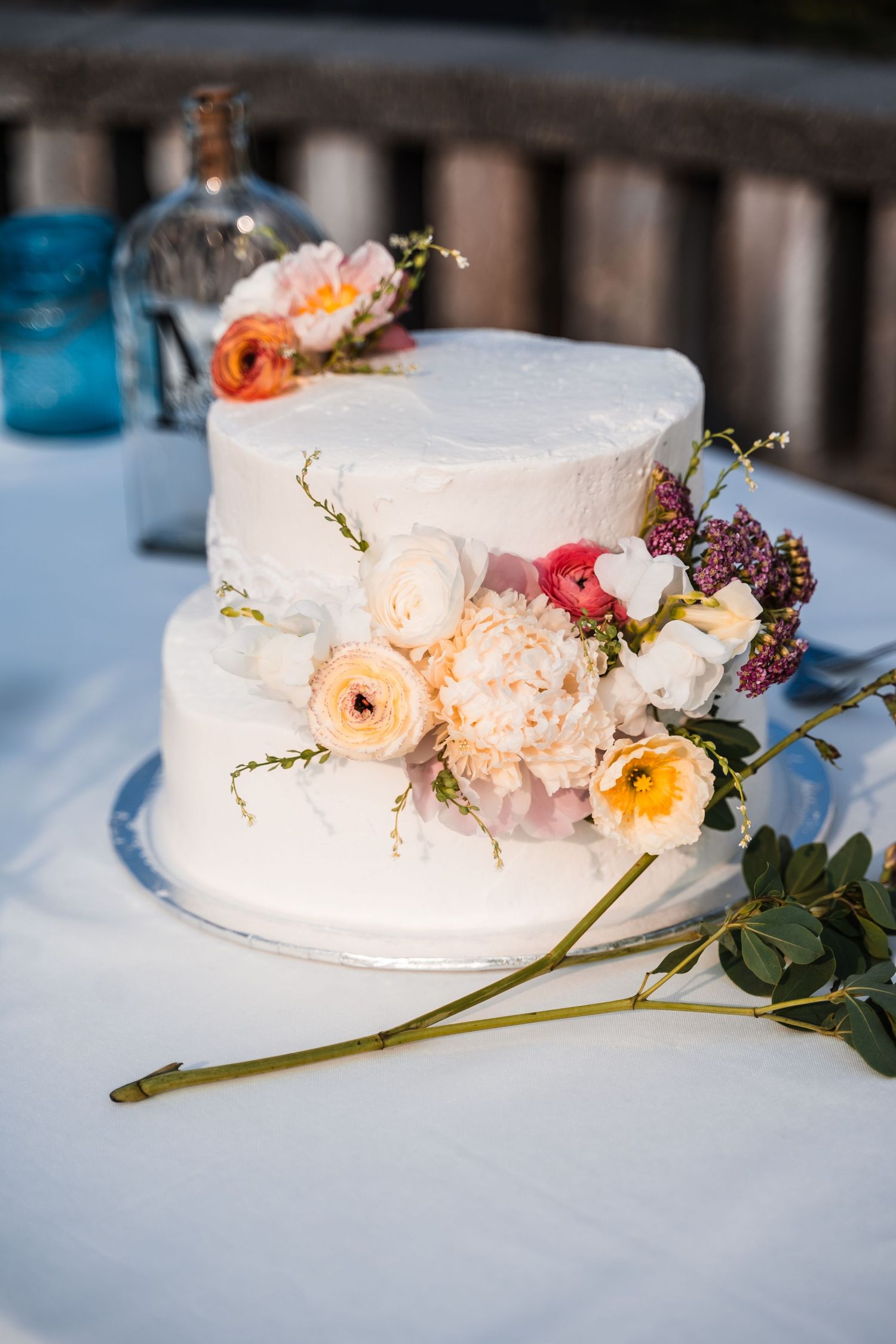 A white frosted wedding cake decorated with fresh peach, yellow and pink flowers sits on a light blue tablecloth.