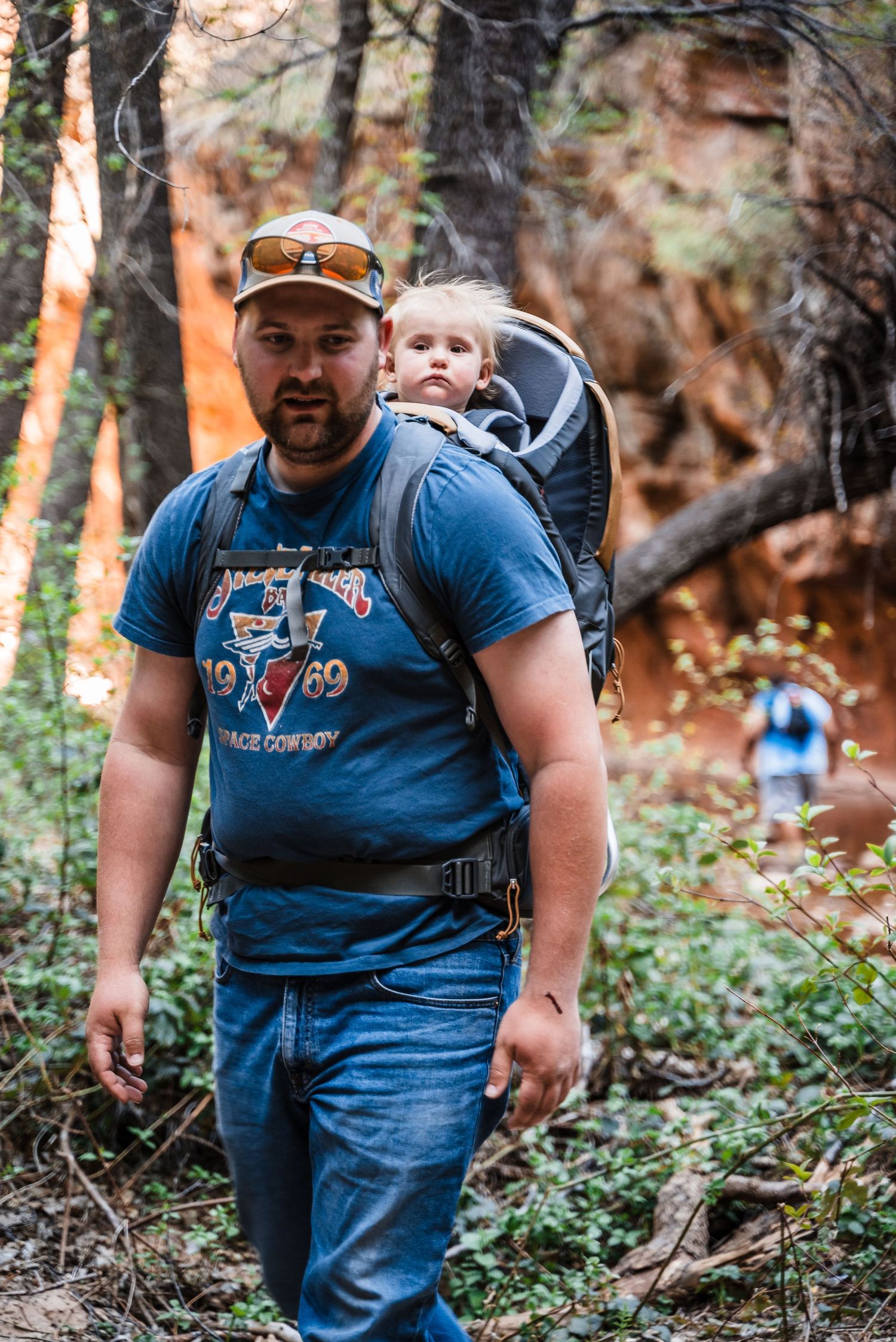 A hiker wearing a blue t-shirt and jeans carries a child in a backpack carrier while walking through a wooded trail.