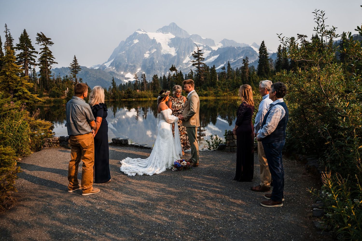 A wedding ceremony takes place in a scenic mountain landscape with evergreen trees and snow-capped peaks at sunset.