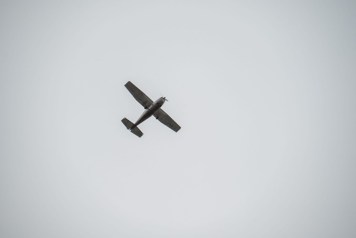 Silhouette of a small propeller aircraft flying against an overcast grey sky.