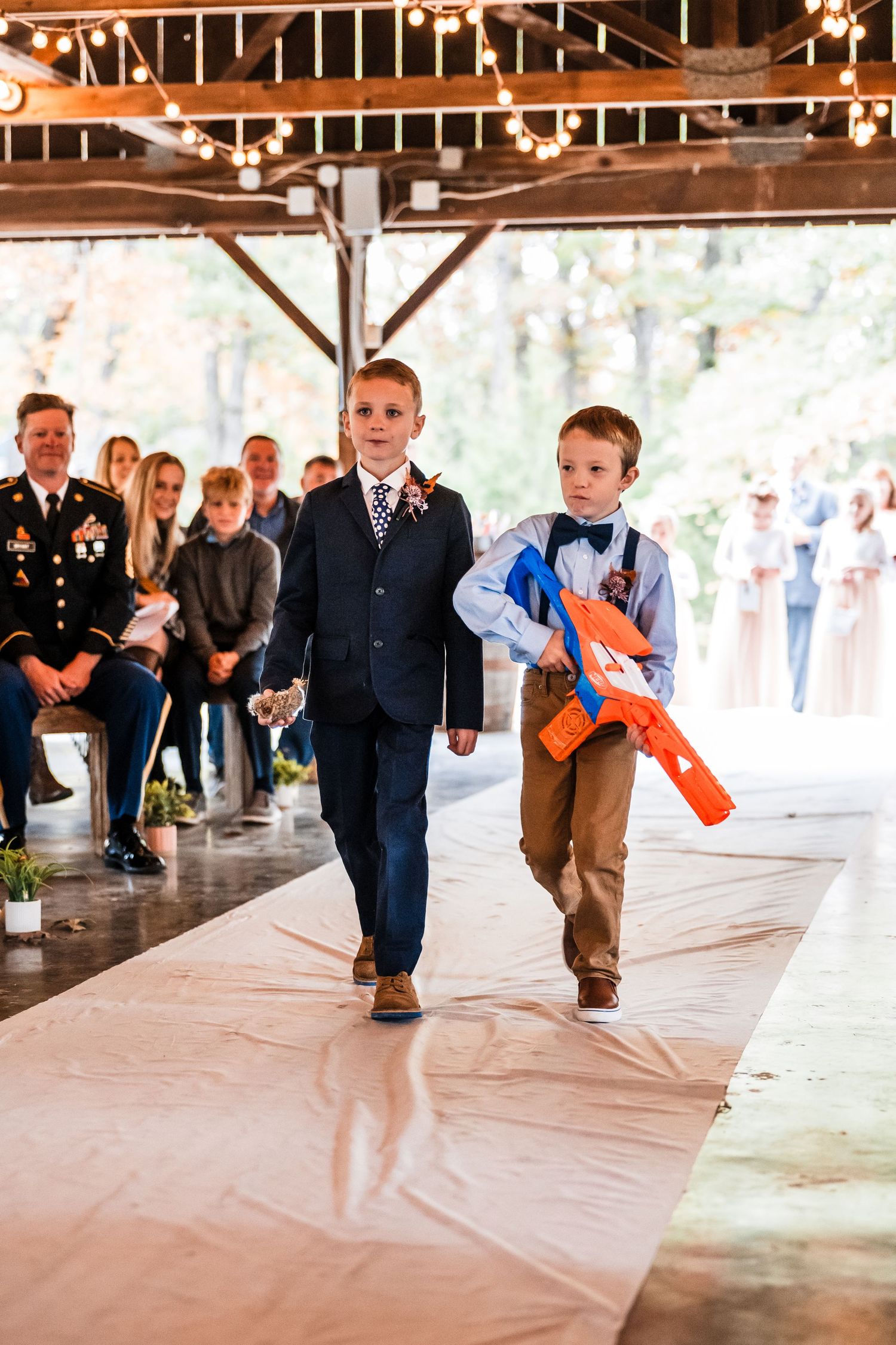Young ring bearers walk down the aisle at a rustic wedding venue decorated with string lights.