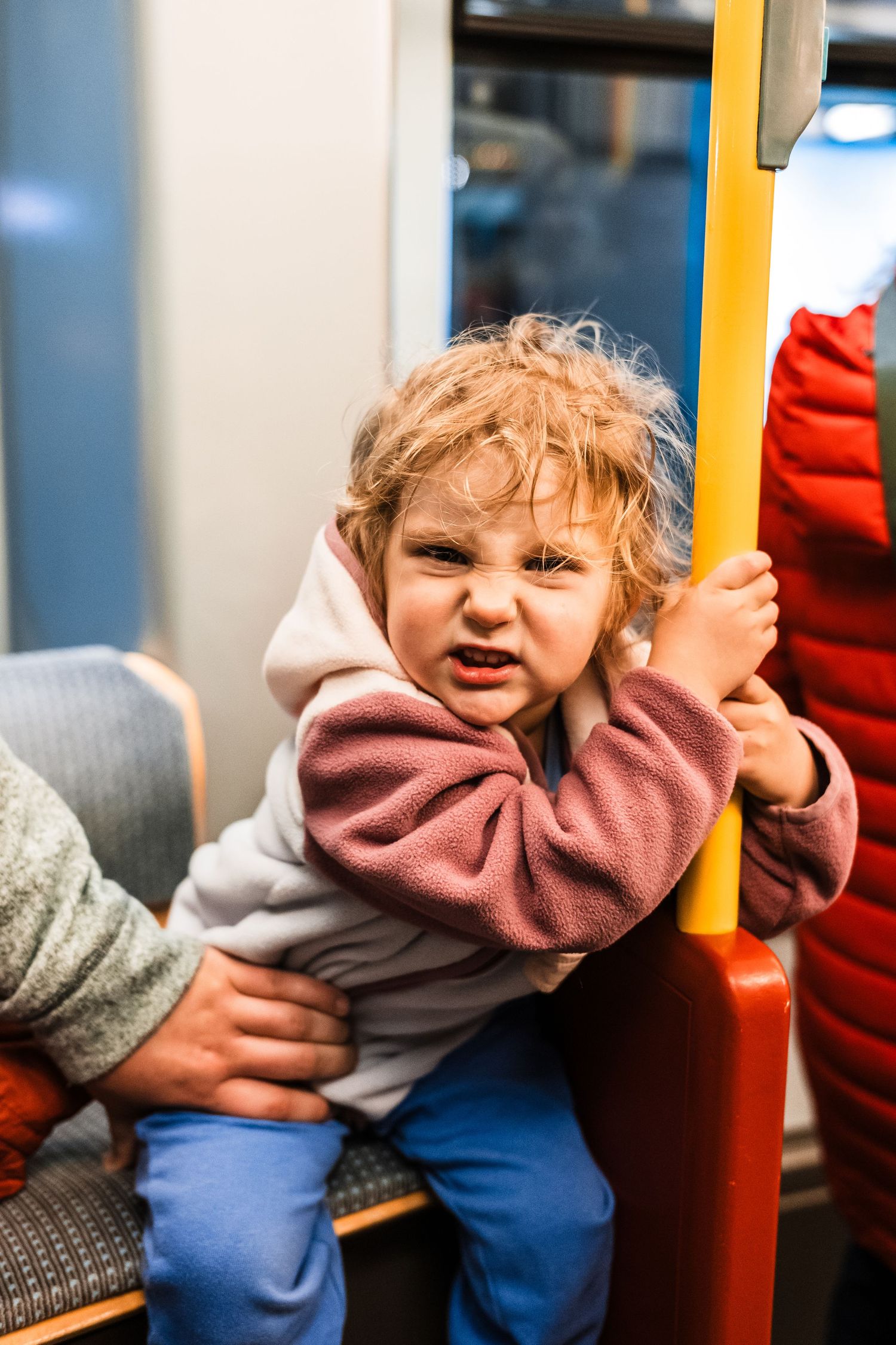 Small child in pink sweater holding yellow pole while riding public transportation.