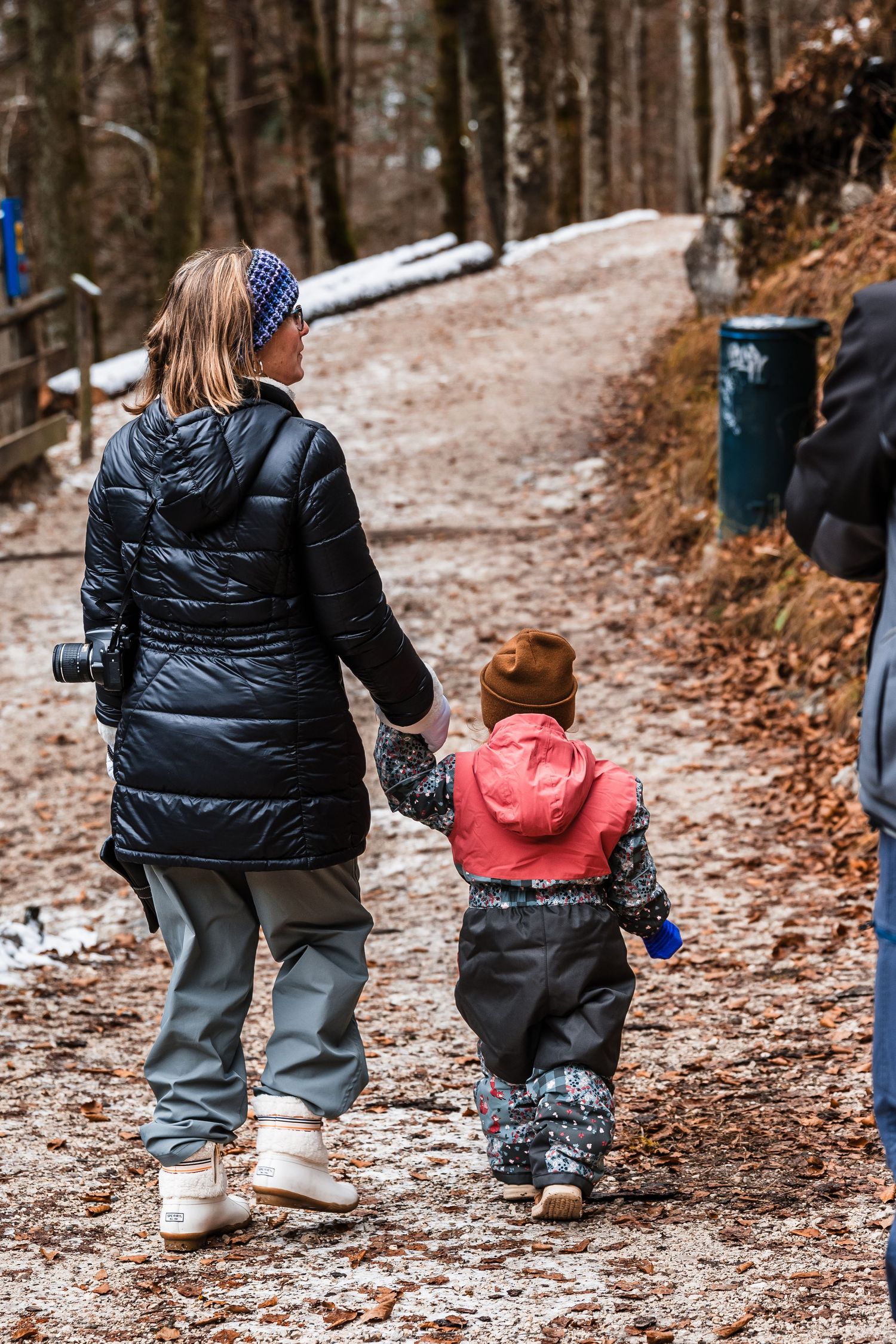 Close-up view of people's legs walking on a forest path covered in light snow and fallen leaves.