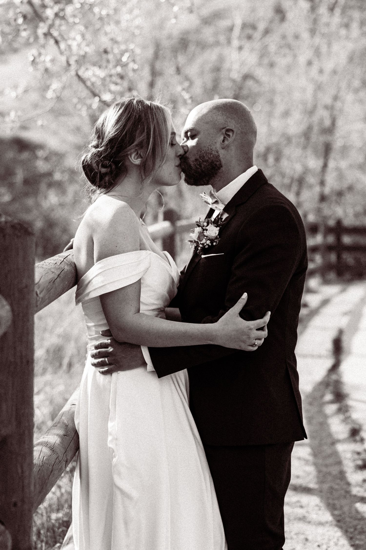 Black and white romantic photo of bride and groom sharing a kiss near a wooden fence in a garden setting.