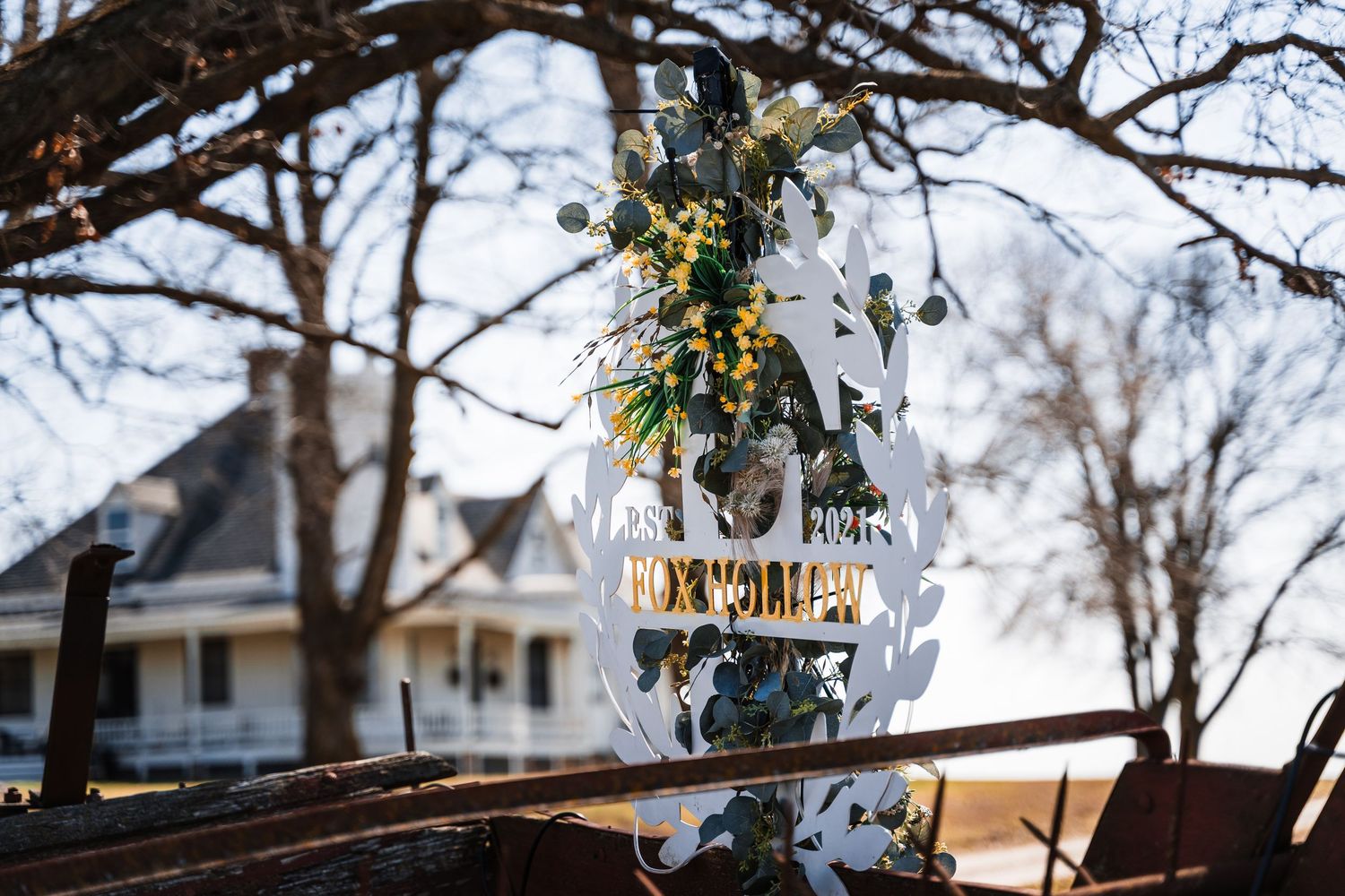 White laser-cut wedding sign adorned with greenery and yellow flowers displayed outdoors among bare winter trees.